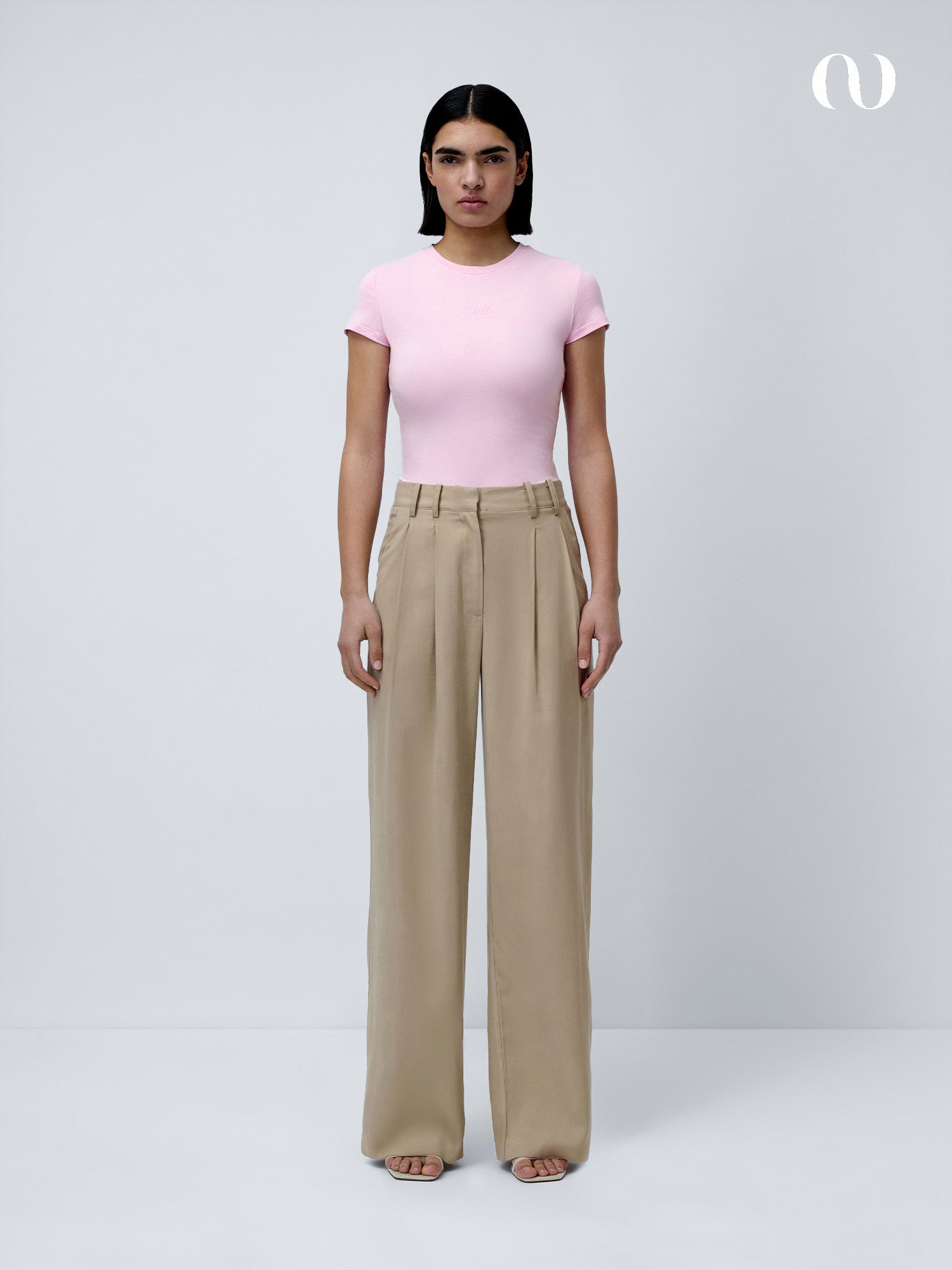 Eine Person steht mit dem Gesicht nach vorne vor einem unifarbenen, hellgrauen Hintergrund und trägt ein rosa Franka T-Shirt von LeGer by Lena Gercke mit kurzen Ärmeln, eine hoch sitzende beige Hose mit weitem Bein und offene Schuhe.
