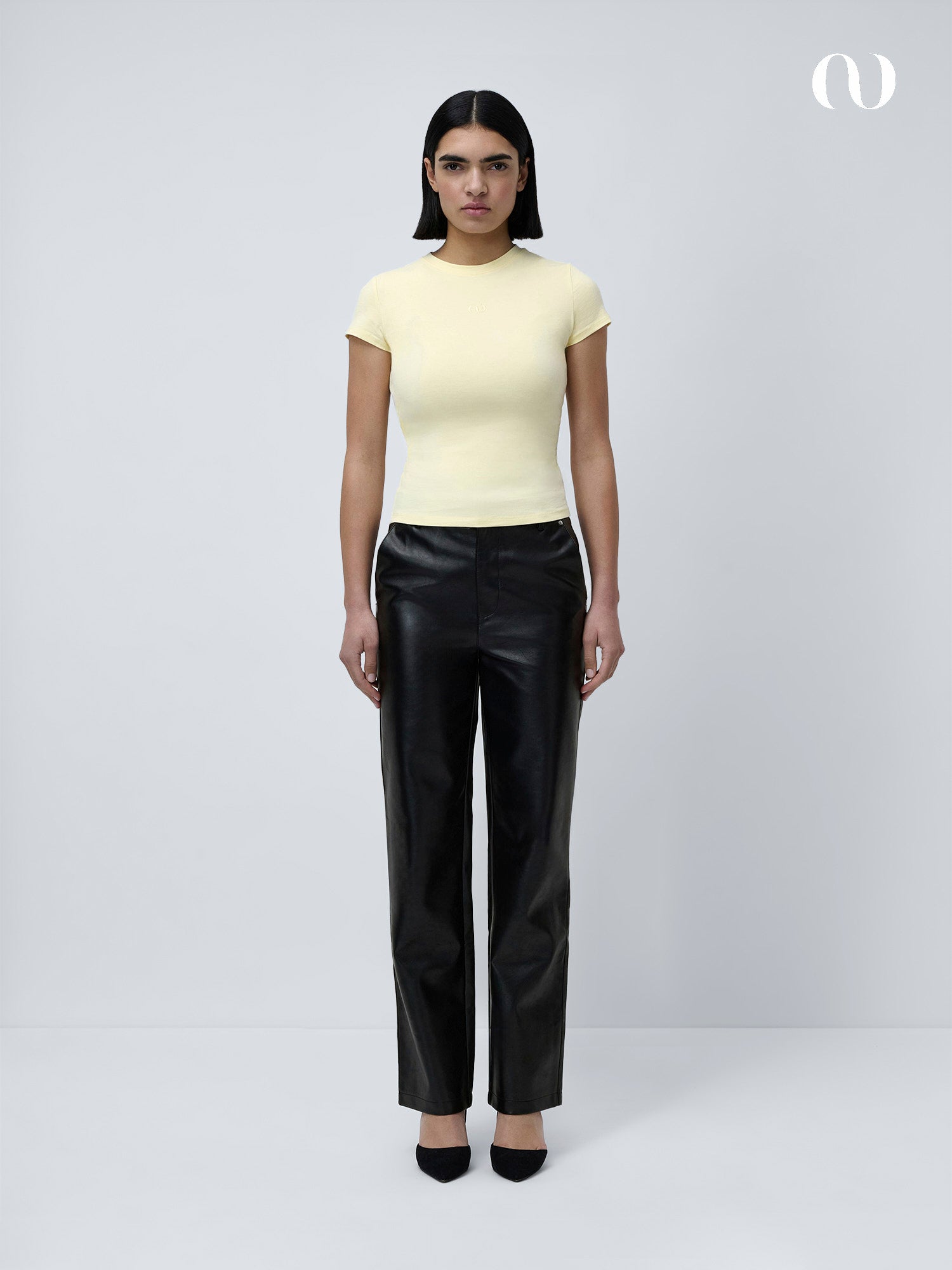 Eine Frau mit glattem dunklem Haar trägt das schmal geschnittene Franka T-Shirt von LeGer by Lena Gercke mit kurzen Ärmeln, gestylt mit schwarzer Lederhose und Absätzen, vor einem schlichten hellen Hintergrund.