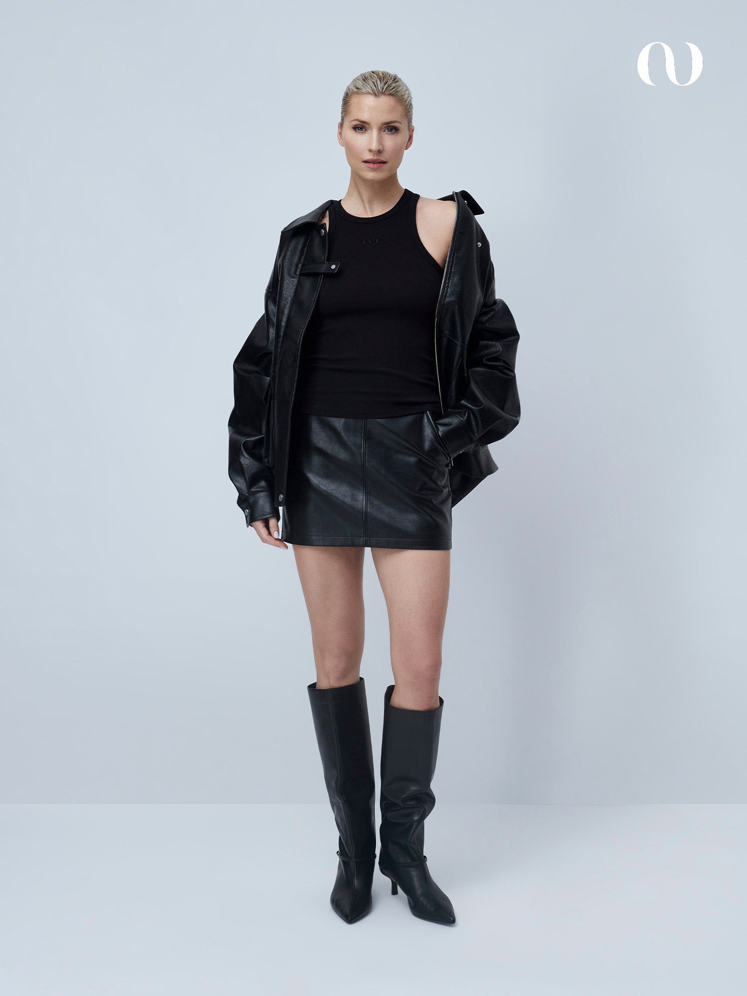 Eine Frau steht vor einem hellgrauen Hintergrund und trägt das schmal geschnittene Dena Top von LeGer by Lena Gercke, einen schwarzen Lederminirock, eine schwarze Lederjacke in Übergröße und kniehohe Stiefel - ihr blondes Haar hat sie für einen selbstbewussten Damen Basic Look zurückgesteckt.