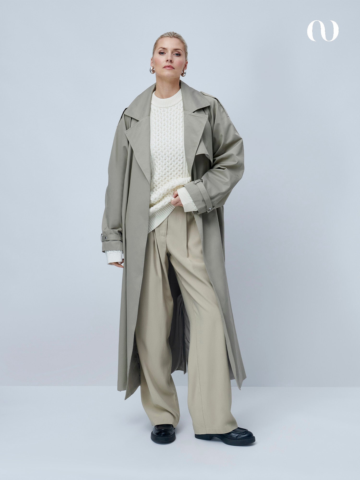 Eine Frau mit kurzen blonden Haaren und Ohrringen steht vor einem hellgrauen Hintergrund und trägt einen langen beigen Trenchcoat, eine beigefarbene Hose mit weitem Bein, schwarze Schuhe und den LeGer by Lena Gercke Danika Jumper, einen übergroßen Zopfstrickpullover mit langen Ärmeln.