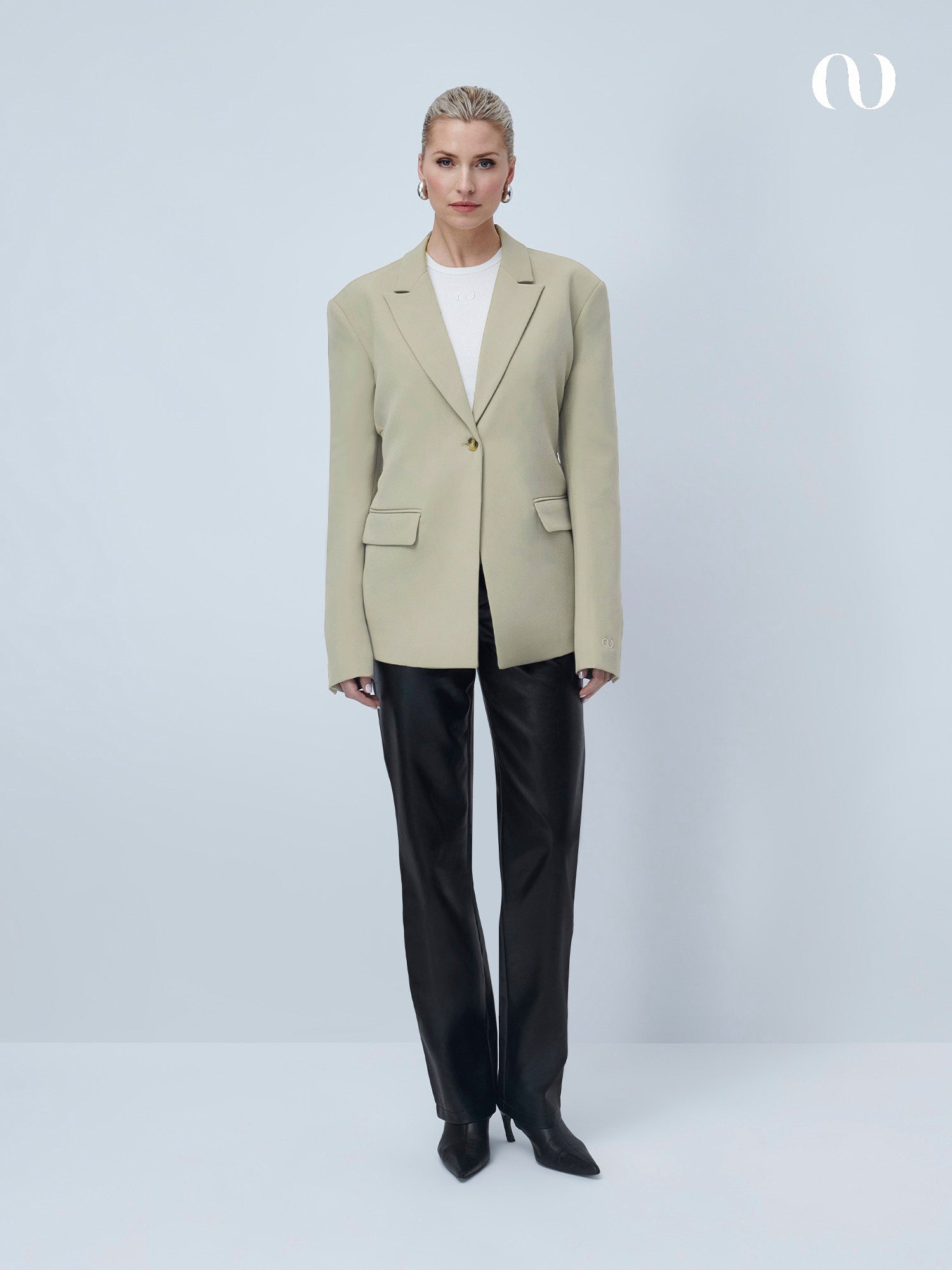 Eine Frau trägt den Line Blazer von LeGer by Lena Gercke in Beige vor grauem Hintergrund, kombiniert mit weißem Top, schwarzen Lederhosen und spitzen schwarzen Schuhen. Sie hat blonde, zurückgebundene Haare und blickt in die Kamera.