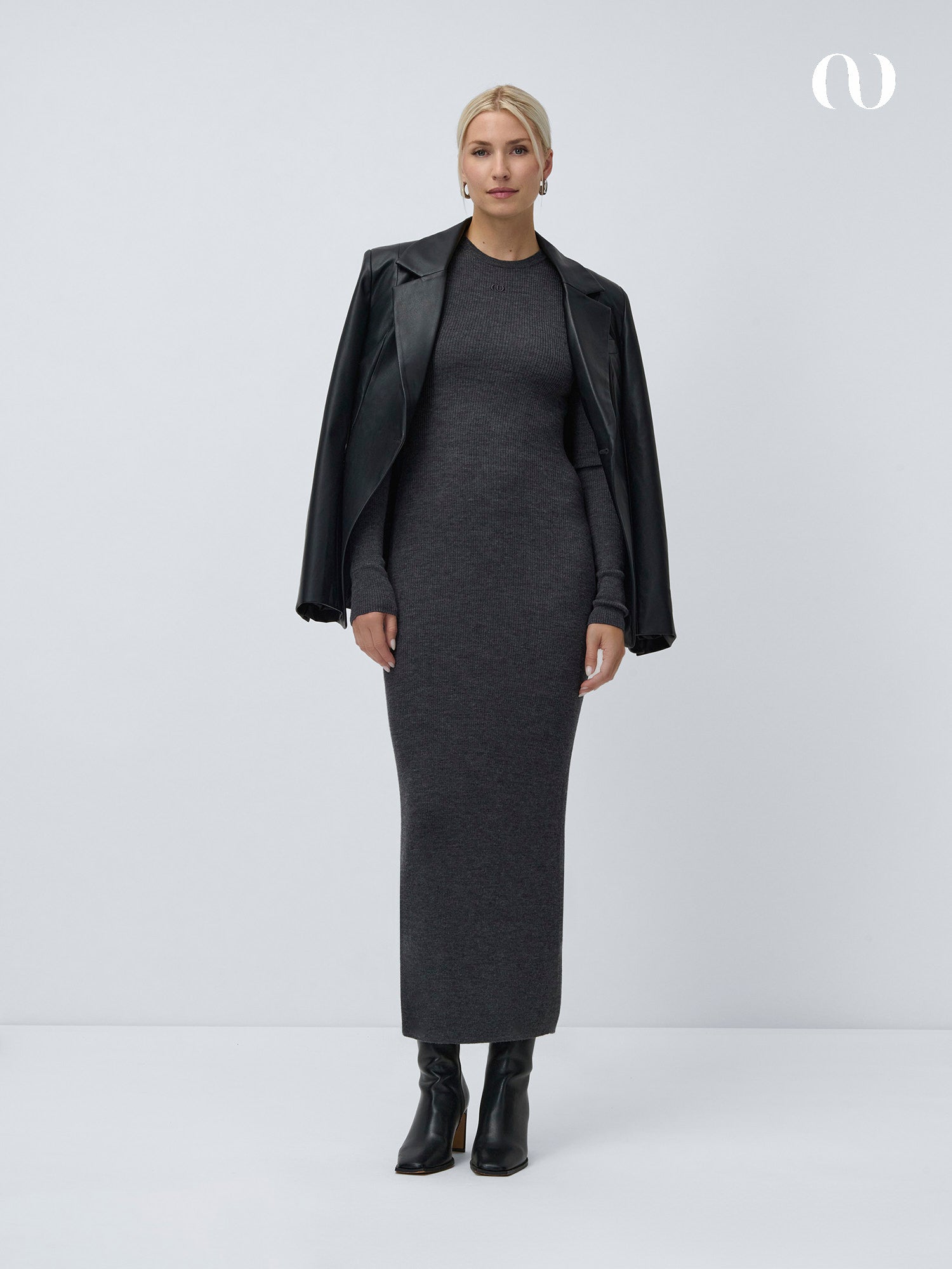 Eine Frau mit blonden Haaren steht vor einem weißen Hintergrund und trägt das Mina Dress von LeGer by Lena Gercke in Dunkelgrau, dazu schwarze Stiefel mit Absatz und eine schwarze Lederjacke mit überlangen Ärmeln, die sie über die Schultern gelegt hat.