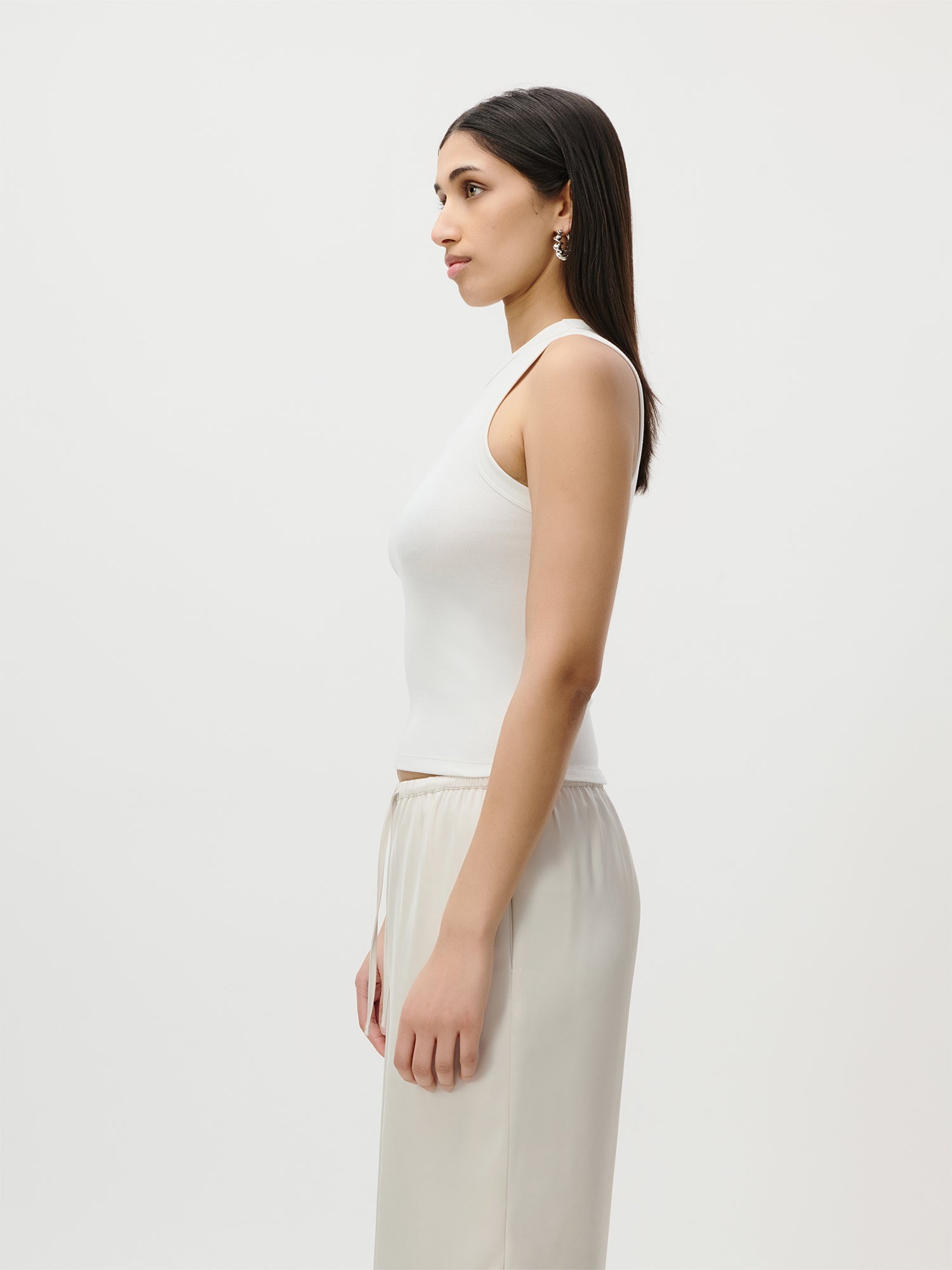 Eine Frau mit glattem, dunklem Haar steht im Profil vor einem schlichten weißen Hintergrund. Sie trägt das ärmellose Top „Ria“ mit feiner Rippenstruktur von LeGer by Lena Gercke sowie eine hellbeige Hose und verkörpert damit perfekt eine moderne Capsule Wardrobe.