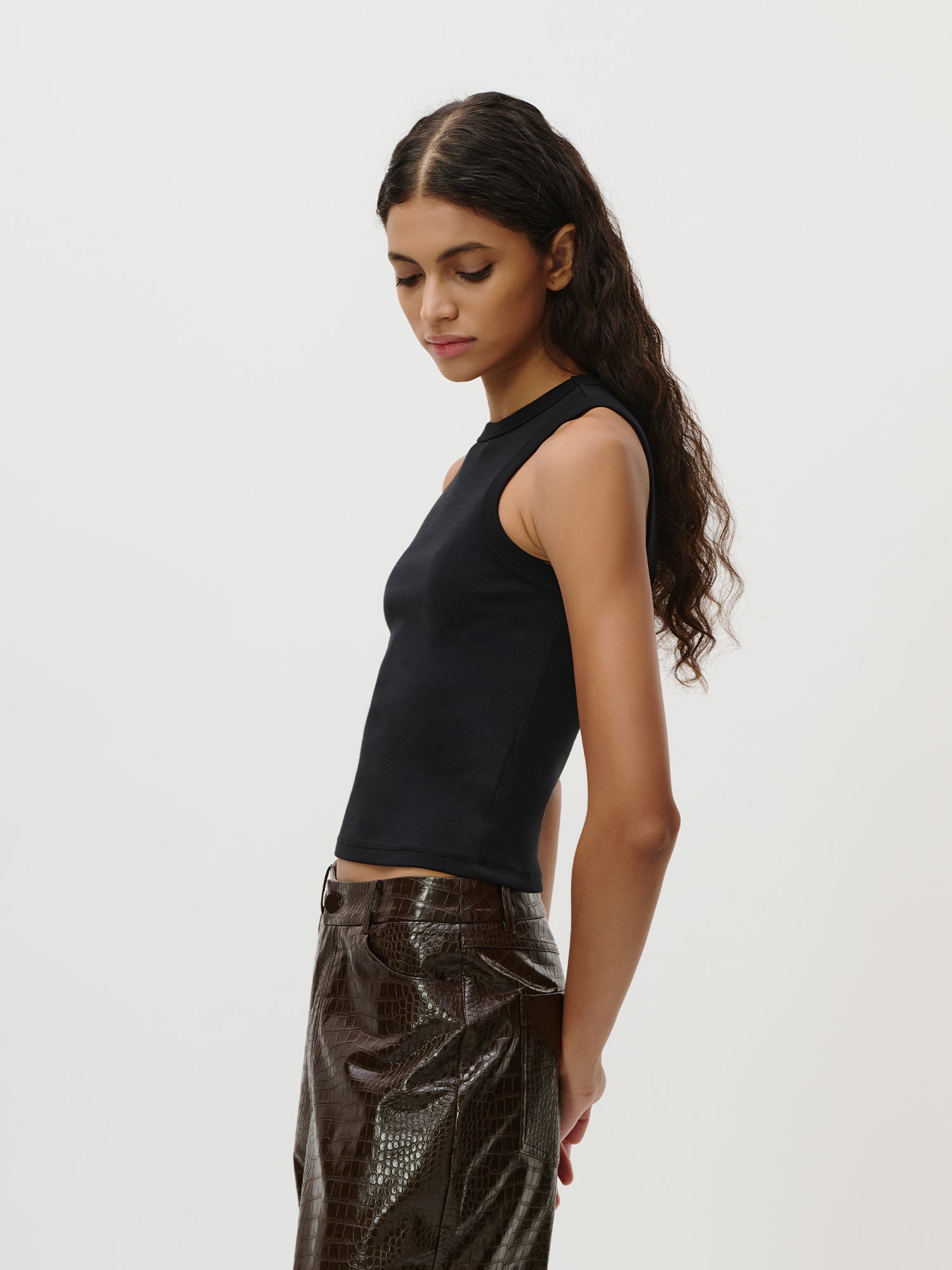 Eine Frau mit langen, welligen Haaren steht im Profil vor einem schlichten Hintergrund. Sie trägt das ärmellose schwarze „Ria“-Top von LeGer by Lena Gercke mit Ripp-Struktur, kombiniert mit einer braunen, strukturierten Hose – ein mühelos schicker Look, der perfekt in jede Capsule Wardrobe passt. Mit ausdruckslosem Gesicht blickt sie nach unten.