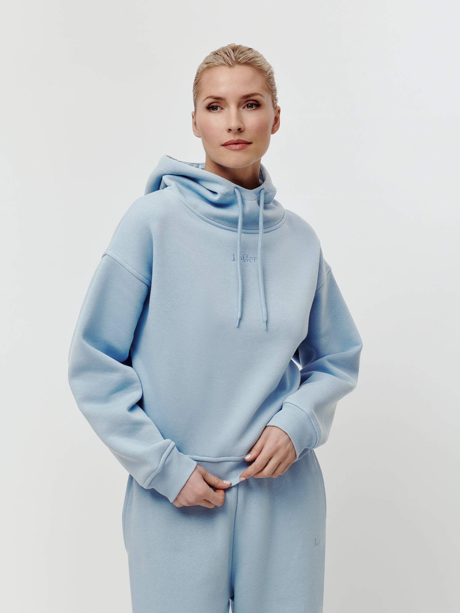 Eine Person mit blonden Haaren, die zu einem Dutt zusammengebunden sind, trägt den hellblauen „Karima“-Hoodie von LeGer by Lena Gercke mit langen Ärmeln und lockerer Passform sowie eine dazu passende Sweathose in normaler Länge. Sie steht vor einem schlichten weißen Hintergrund und blickt selbstbewusst in die Kamera.