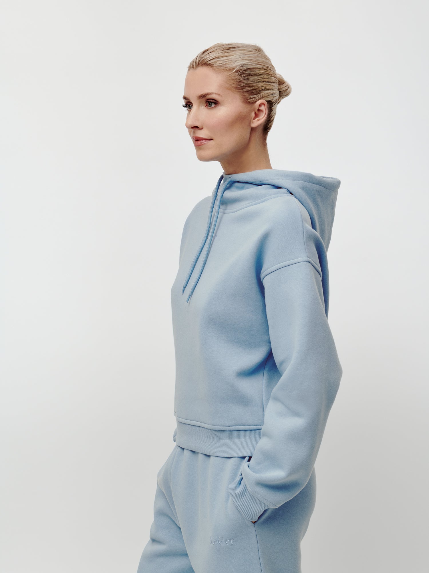 Eine Frau mit blonden Haaren, die zu einem ordentlichen Dutt zusammengebunden sind, trägt den hellblauen „Karima“-Langarm-Hoodie von LeGer by Lena Gercke sowie eine dazu passende Jogginghose in normaler Länge und lockerer Passform. Sie steht vor einem schlichten weißen Hintergrund und blickt nach links.