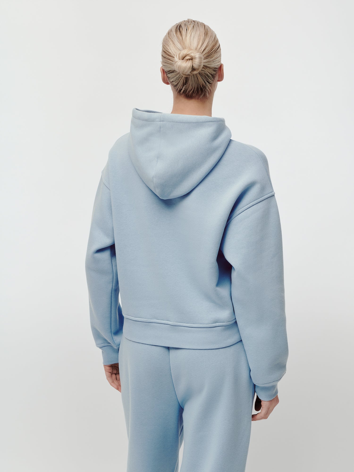 Eine Person mit blonden Haaren, die zu einem ordentlichen Dutt zusammengebunden sind, trägt den hellblauen „Karima“-Hoodie von LeGer by Lena Gercke in lockerer Passform sowie eine dazu passende Jogginghose und steht mit dem Rücken zum Betrachter vor einem schlichten weißen Hintergrund. Der Hoodie hat lange Ärmel und eine normale Länge und bietet sowohl Komfort als auch einen lässigen Stil.
