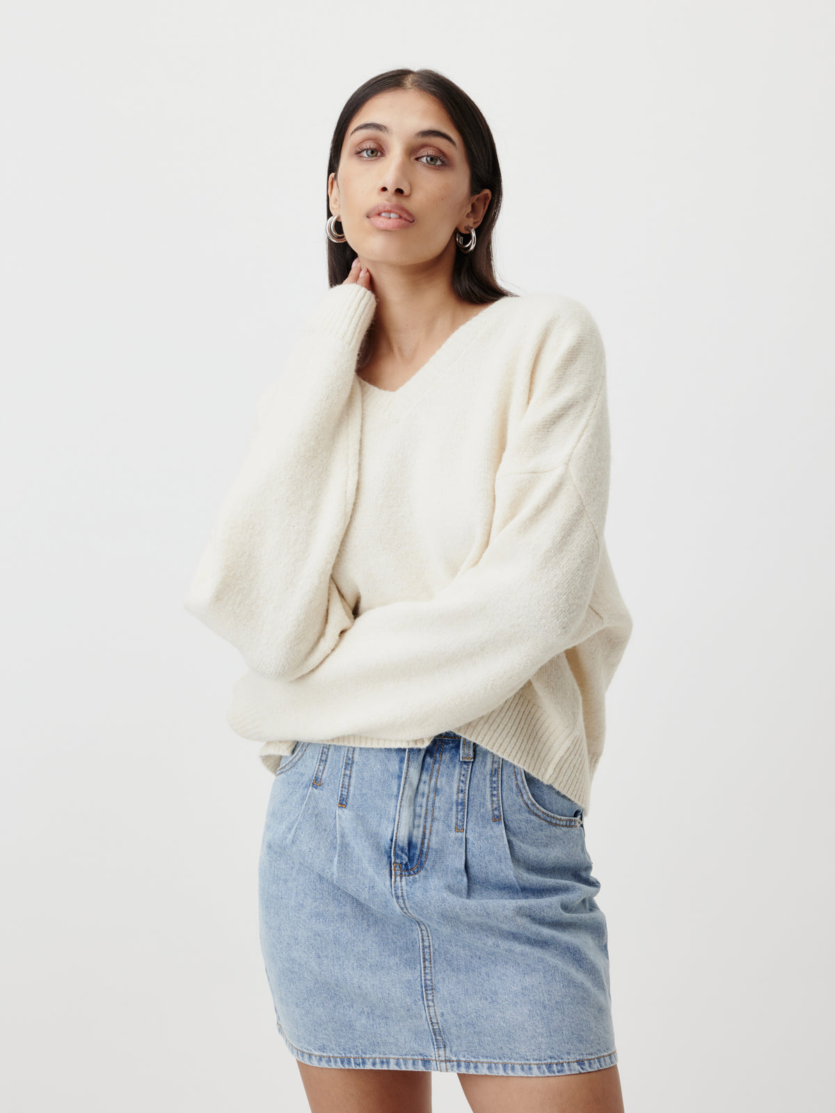 Eine Frau, die den Pullover „Halina“ von LeGer by Lena Gercke aus lockerem weißem Strick mit V-Ausschnitt und einen hellblauen Jeansrock trägt, steht vor einem schlichten weißen Hintergrund und blickt selbstbewusst in die Kamera, während sie mit einer Hand ihr Gesicht berührt.
