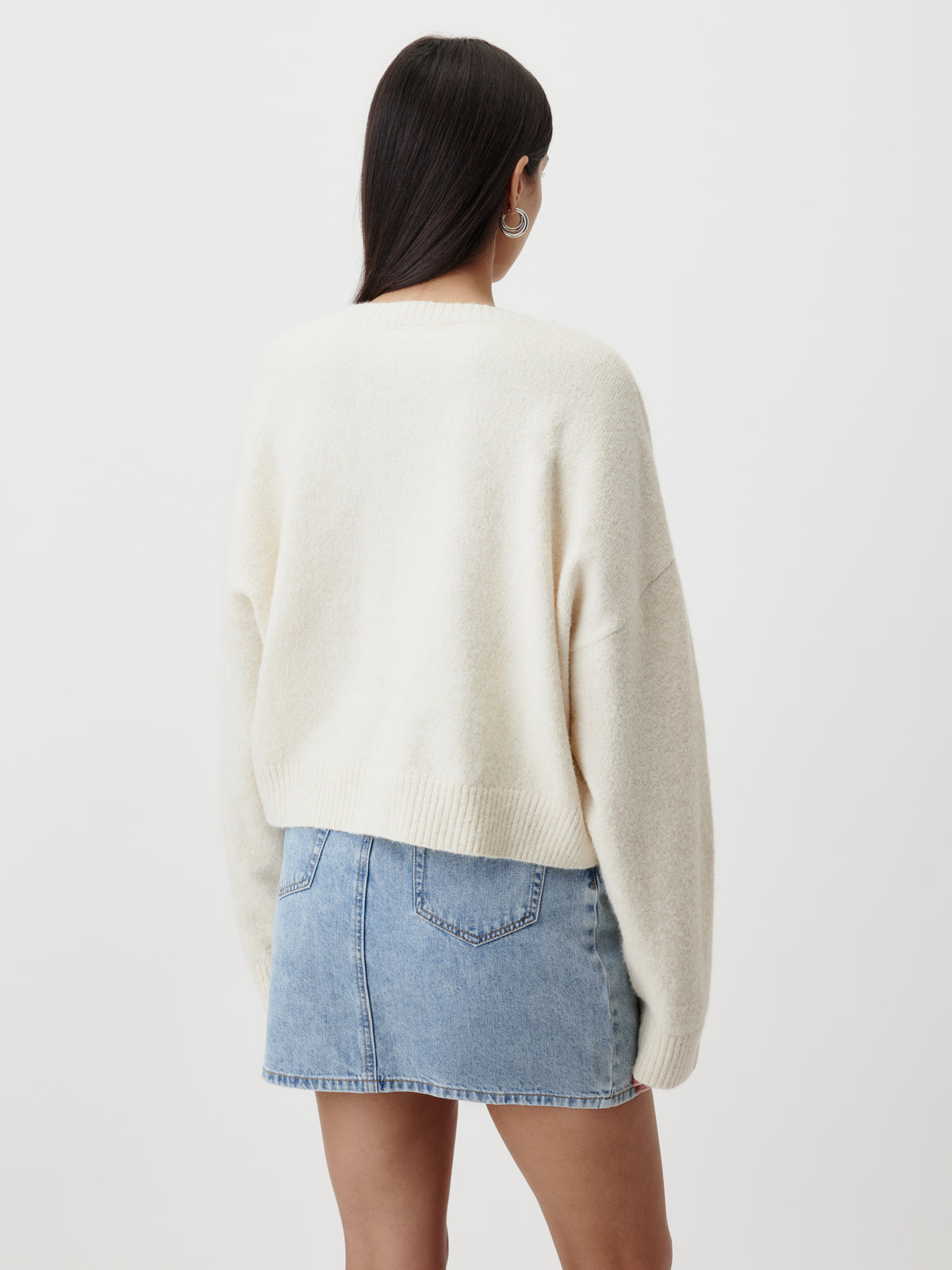 Eine Person mit langen, dunklen Haaren trägt einen übergroßen, cremefarbenen „Halina“-Pullover von LeGer by Lena Gercke und einen hellblauen Jeansrock. Sie steht mit dem Rücken zur Kamera vor einem schlichten weißen Hintergrund.