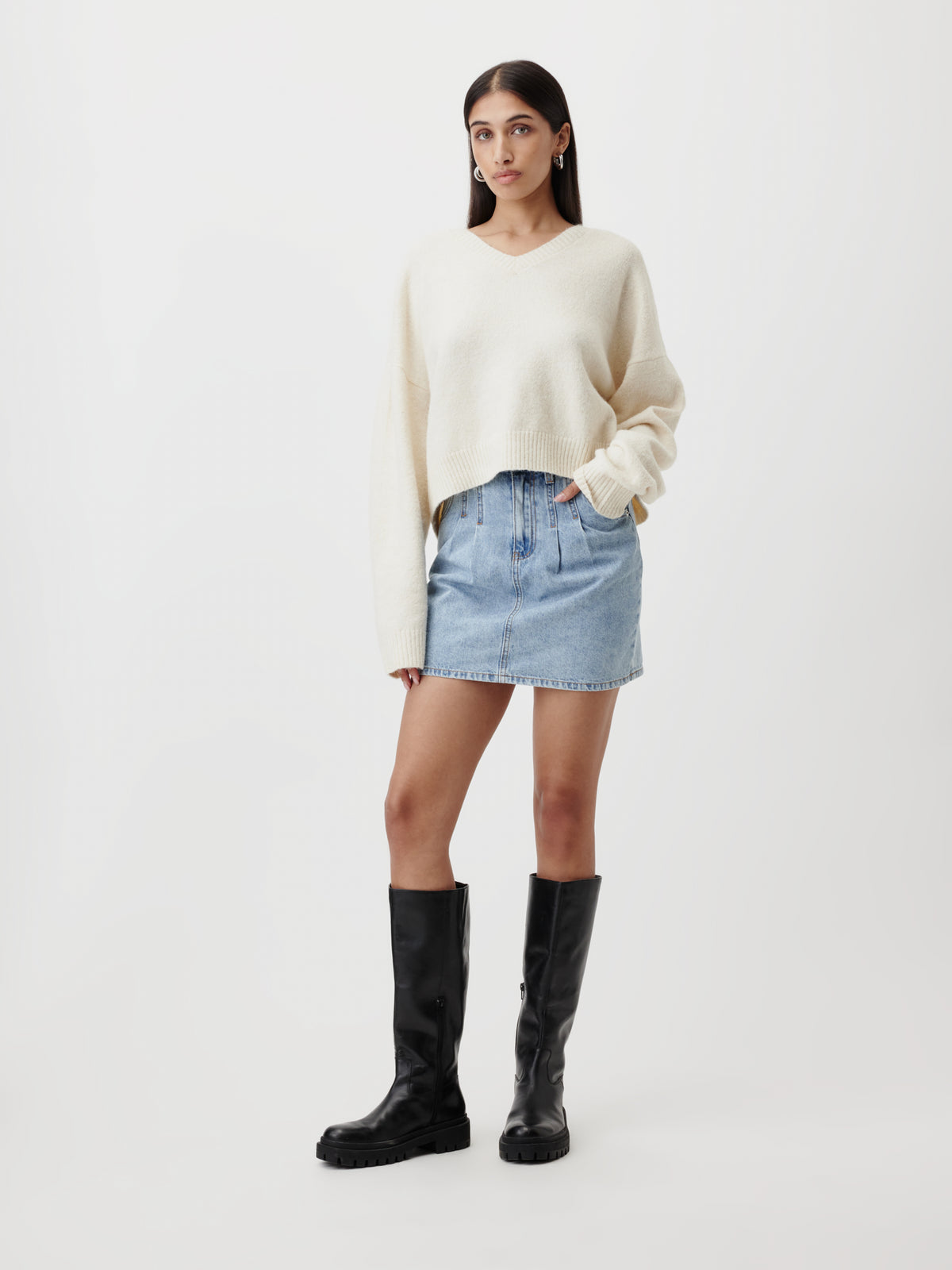 Vor einem weißen Hintergrund steht eine Frau, die den übergroßen, cremefarbenen „Halina“-Pullover von LeGer by Lena Gercke trägt, kombiniert mit einem hellen Jeans-Minirock und schwarzen kniehohen Stiefeln. Sie hat langes braunes Haar und einen ausdruckslosen Gesichtsausdruck, wobei sie eine Hand in die Rocktasche gesteckt hat.