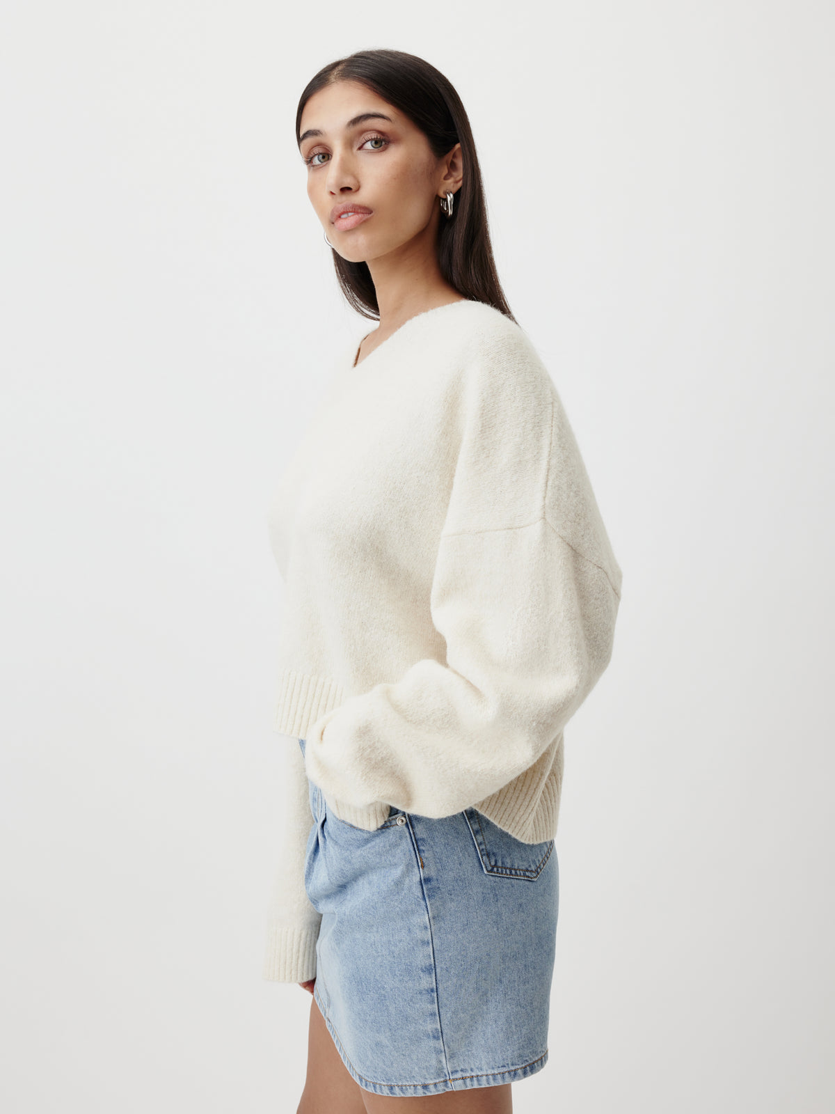 Eine Frau steht im Profil und trägt den cremefarbenen Pullover „Halina“ von LeGer by Lena Gercke, der sich durch einen Oversize-Schnitt und einen V-Ausschnitt auszeichnet, dazu einen hellblauen Jeansrock. Sie hat langes dunkles Haar, ist nur dezent geschminkt und hat einen neutralen Gesichtsausdruck. Der Hintergrund ist schlicht weiß.