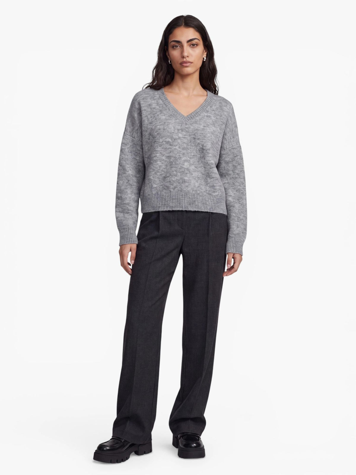 Eine Person steht mit dem Blick nach vorne und trägt den übergroßen, hellgrauen „Halina“-Pullover von LeGer by Lena Gercke, kombiniert mit einer dunkelgrauen Hose und schwarzen, klobigen Schuhen, vor einem schlichten weißen Hintergrund.