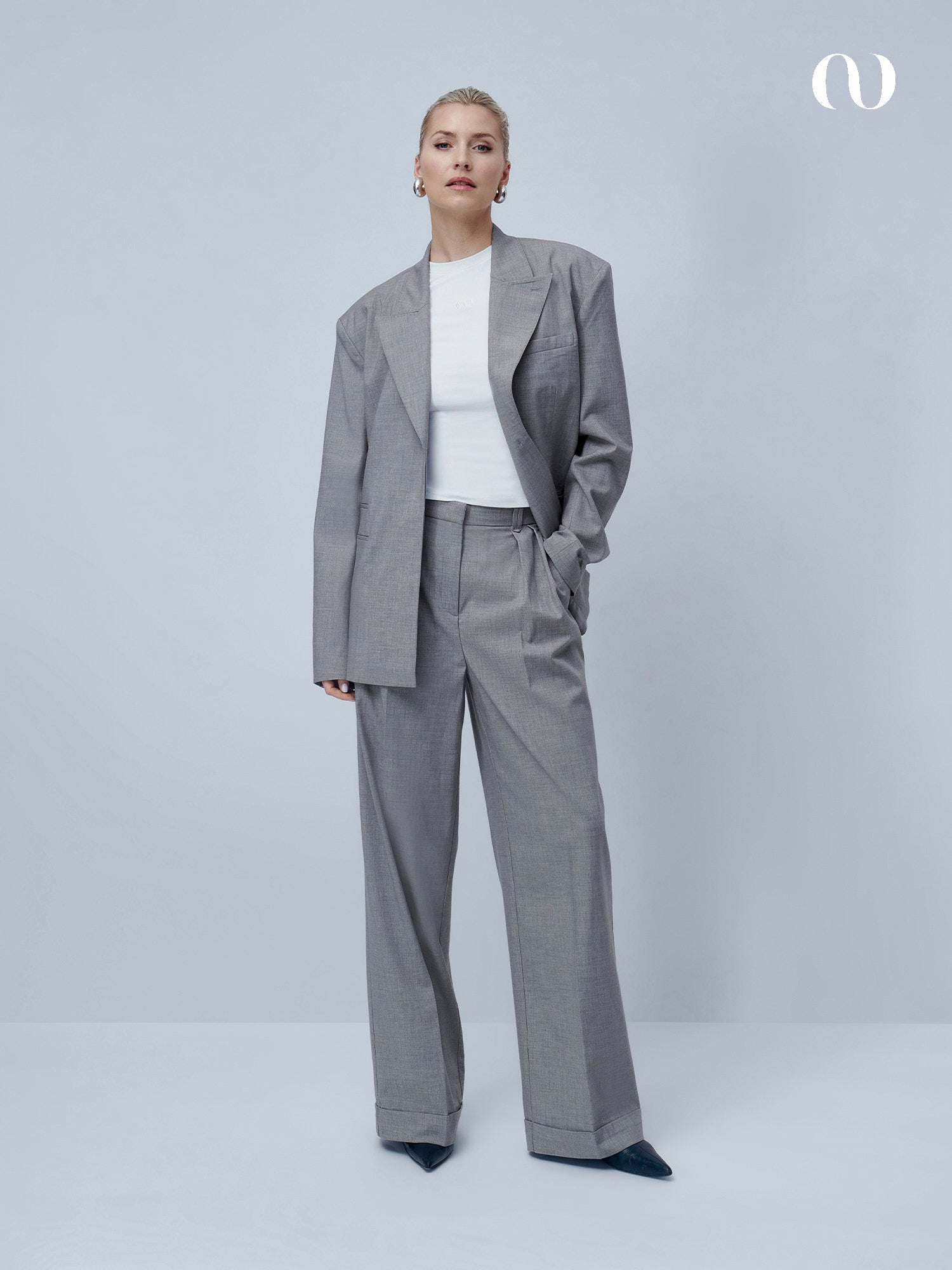 Eine Frau mit kurzen blonden Haaren posiert selbstbewusst vor einem hellgrauen Hintergrund. Sie trägt die Jale Pants Tall von LeGer by Lena Gercke in Grau mit hoher Taille und Bügelfalten, kombiniert mit einem weißen T-Shirt und schwarzen spitzen Schuhen.