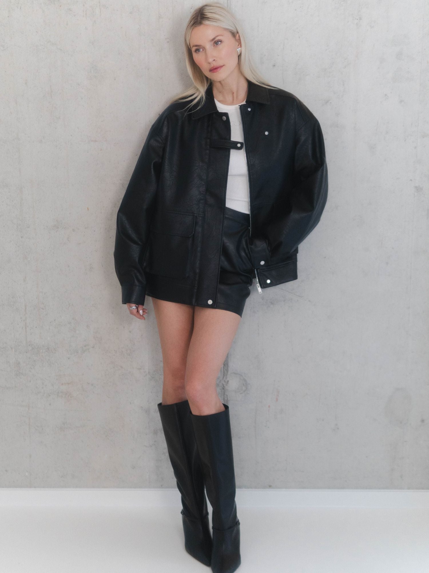 Eine Frau mit langen blonden Haaren steht vor einer hellgrauen Wand und trägt den Ella Skirt von LeGer by Lena Gercke - eine schwarze Lederjacke in Übergröße, ein schwarzer Ella Skirt mit hoher Taille, ein weißes Top und schwarze kniehohe Stiefel vervollständigen ihren Look.