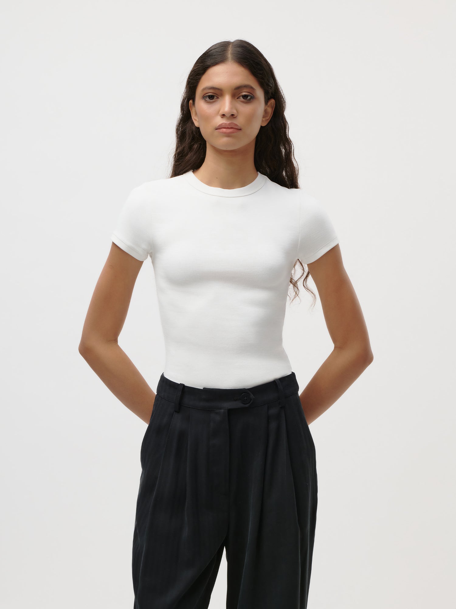 Eine Frau mit langen, gewellten Haaren trägt ein figurbetontes „Ria“-Jersey-Kurzarm-T-Shirt von LeGer by Lena Gercke sowie eine schwarze Hose mit hoher Taille und steht mit hinter dem Rücken verschränkten Händen vor einem schlichten weißen Hintergrund.