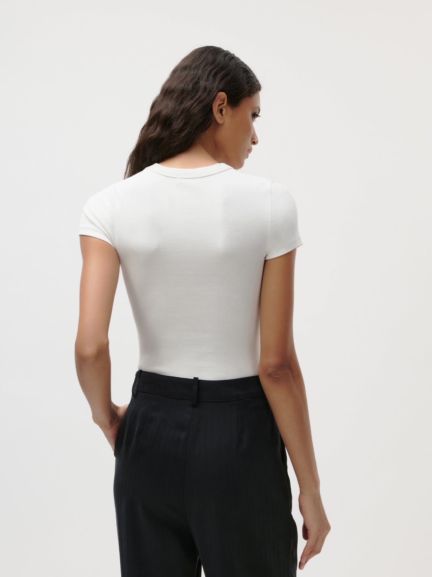 Eine Frau steht mit dem Rücken zur Kamera und trägt ein eng anliegendes weißes „Ria“-T-Shirt von LeGer by Lena Gercke im Jersey-Kurzarm-Stil sowie eine schwarze Hose mit hoher Taille vor einem schlichten, hellen Hintergrund.