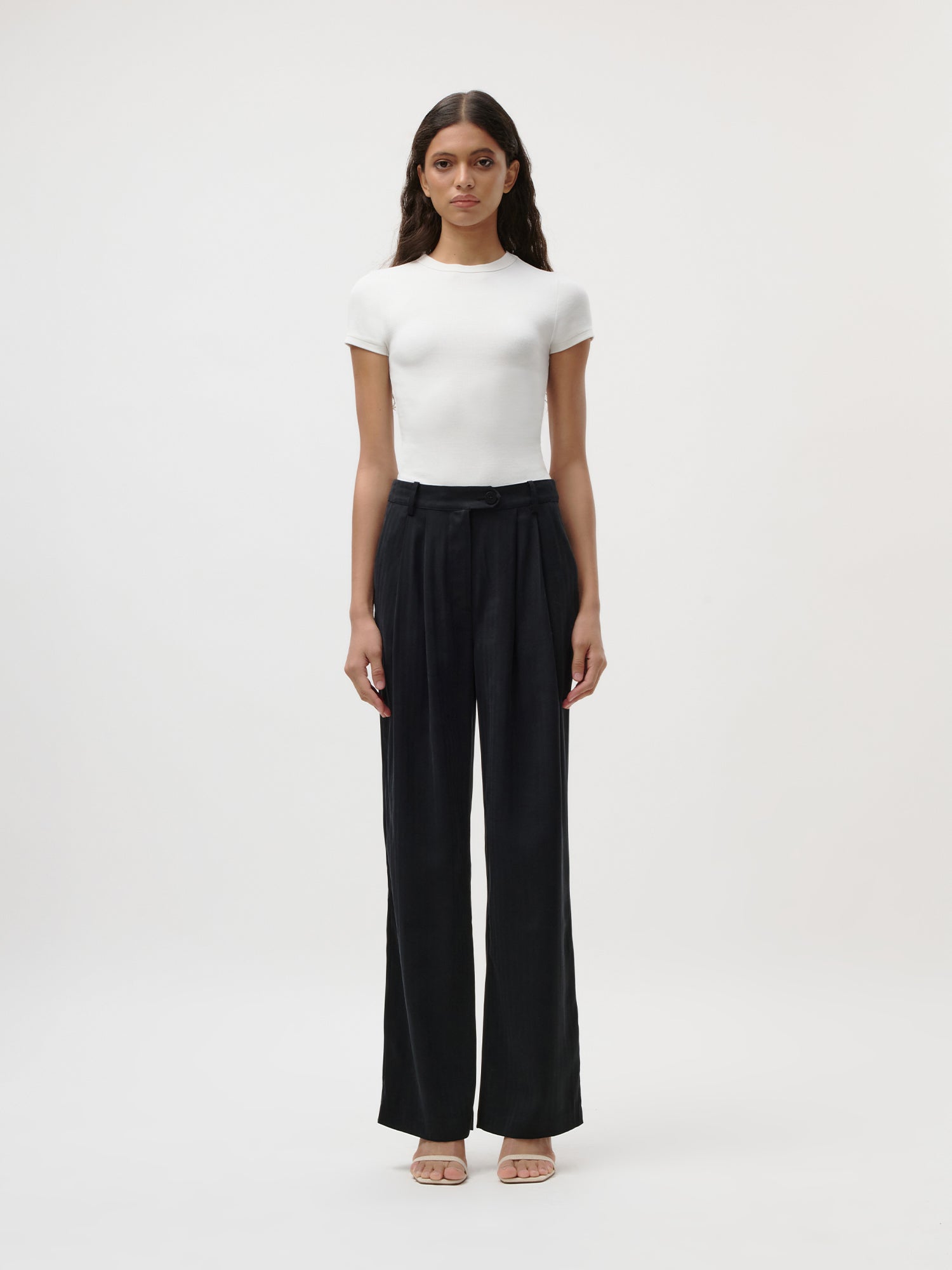 Eine Frau steht mit dem Blick nach vorne vor einem schlichten weißen Hintergrund. Sie trägt ein eng anliegendes weißes „Ria“-T-Shirt von LeGer by Lena Gercke, eine schwarze Hose mit hoher Taille und weitem Bein sowie hautfarbene Pumps mit offener Spitze.