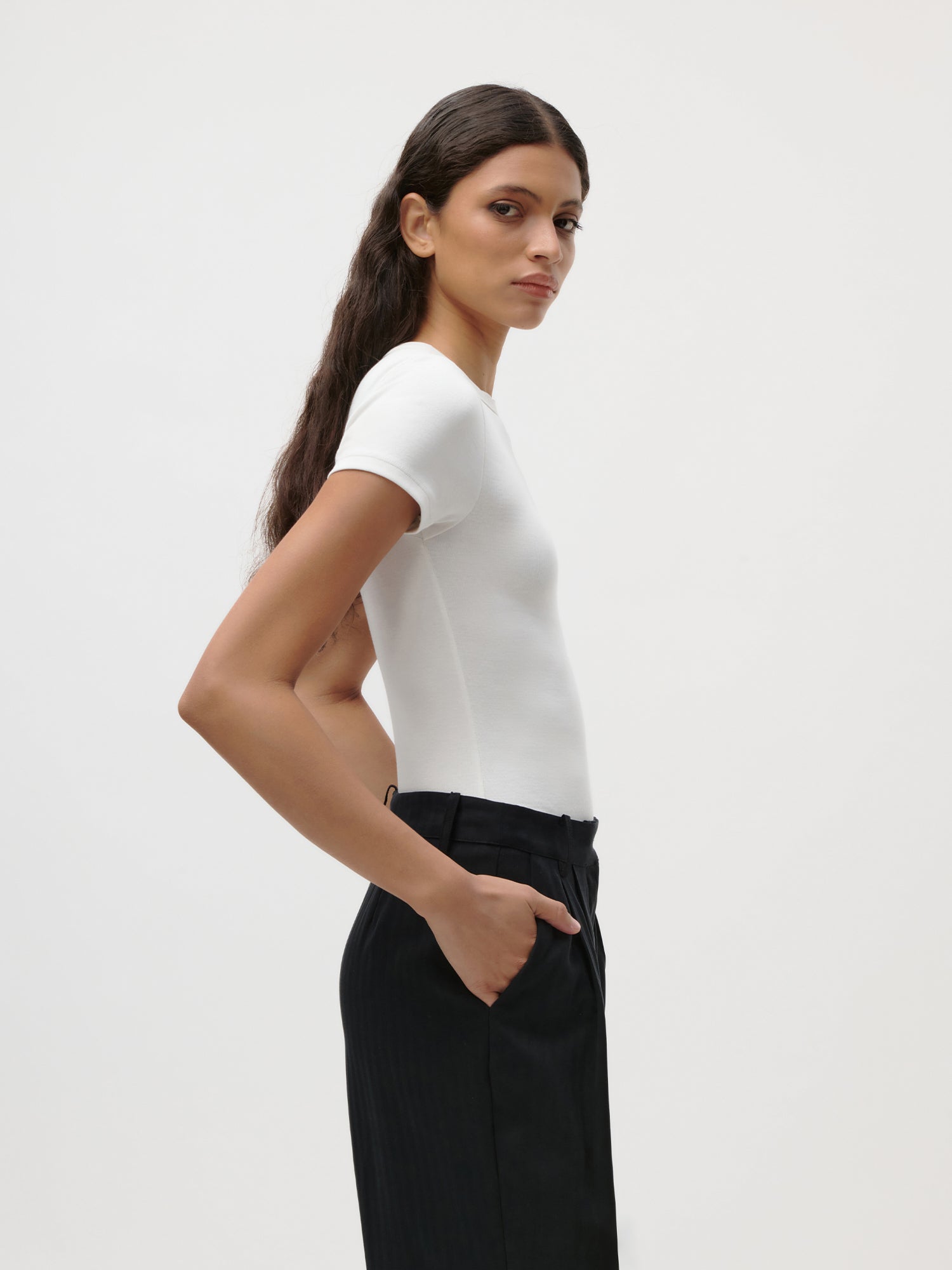 Vor einem schlichten, hellen Hintergrund steht eine Frau mit langen braunen Haaren im Profil, eine Hand in der Tasche. Sie trägt das figurbetonte, weiße „Ria“-T-Shirt von LeGer by Lena Gercke mit kurzen Ärmeln, dazu eine schwarze Hose.