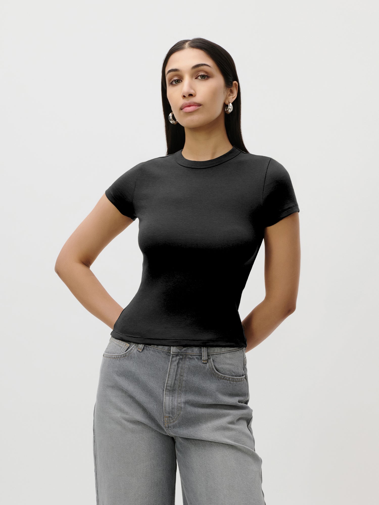 Eine Frau steht vor einem schlichten Hintergrund und trägt das eng anliegende schwarze „Ria“-T-Shirt von LeGer by Lena Gercke aus Jersey mit kurzen Ärmeln sowie hellgraue Jeans; ihre Hände ruhen auf den Hüften. Sie hat langes dunkles Haar und trägt Ohrringe.