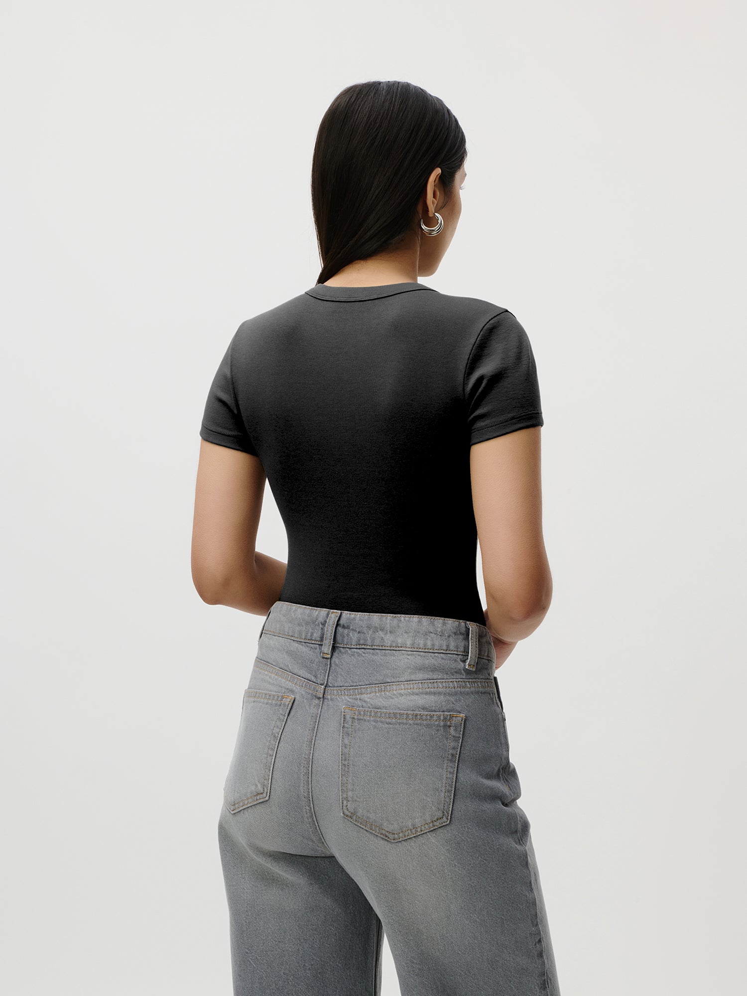 Eine Frau mit langen dunklen Haaren, die das schwarze „Ria“-Slim-Fit-T-Shirt von LeGer by Lena Gercke und eine hellblaue Jeans trägt, steht mit dem Rücken zur Kamera vor einem schlichten hellen Hintergrund.