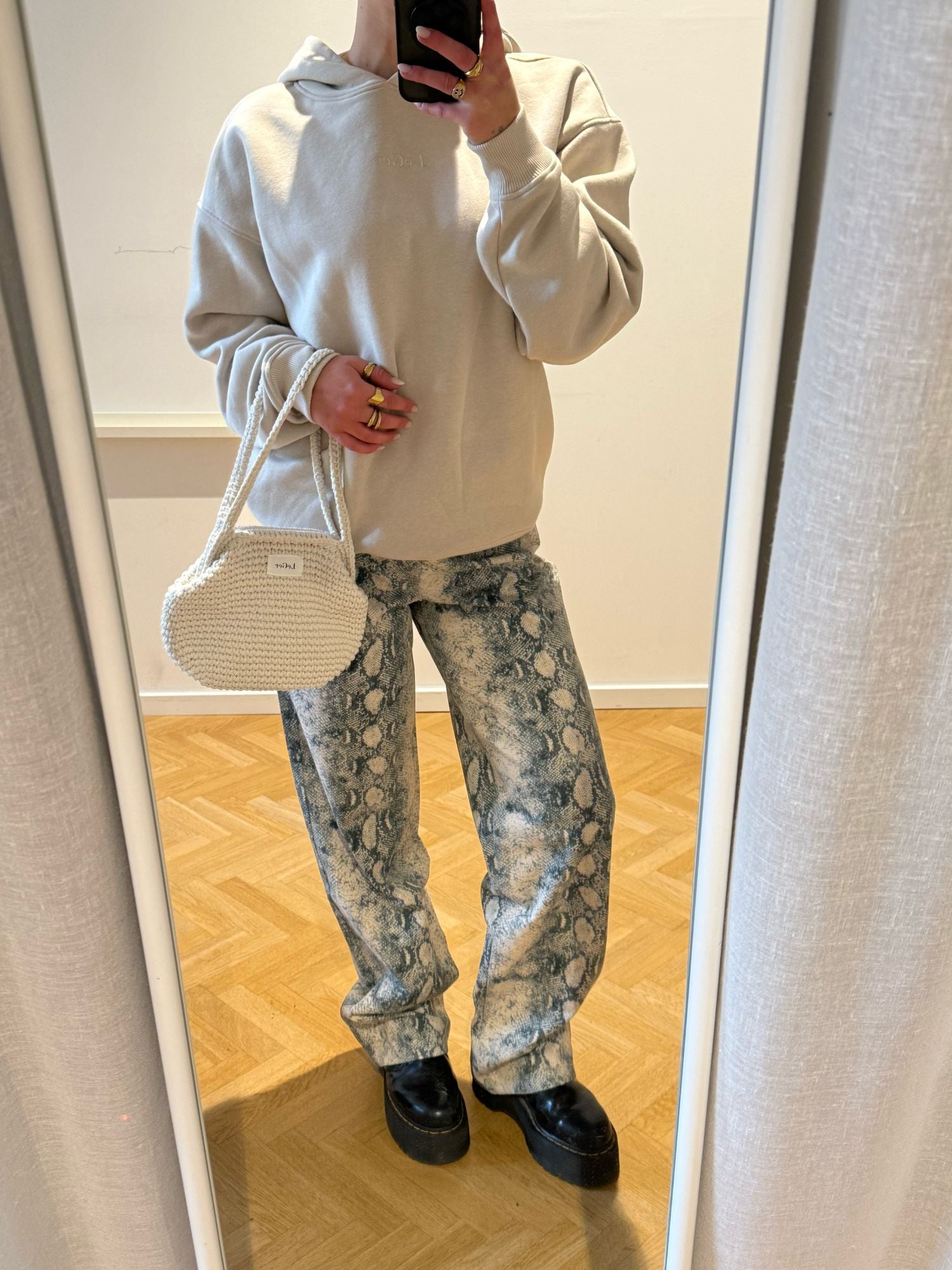 Person, die drinnen auf einem hellen Holzboden ein Spiegel-Selfie macht. Sie trägt einen beigen Kapuzenpulli, die Cleo Pants Tall in Grau und Weiß von LeGer by Lena Gercke, klobige schwarze Schuhe und eine kleine weiße, strukturierte Handtasche.