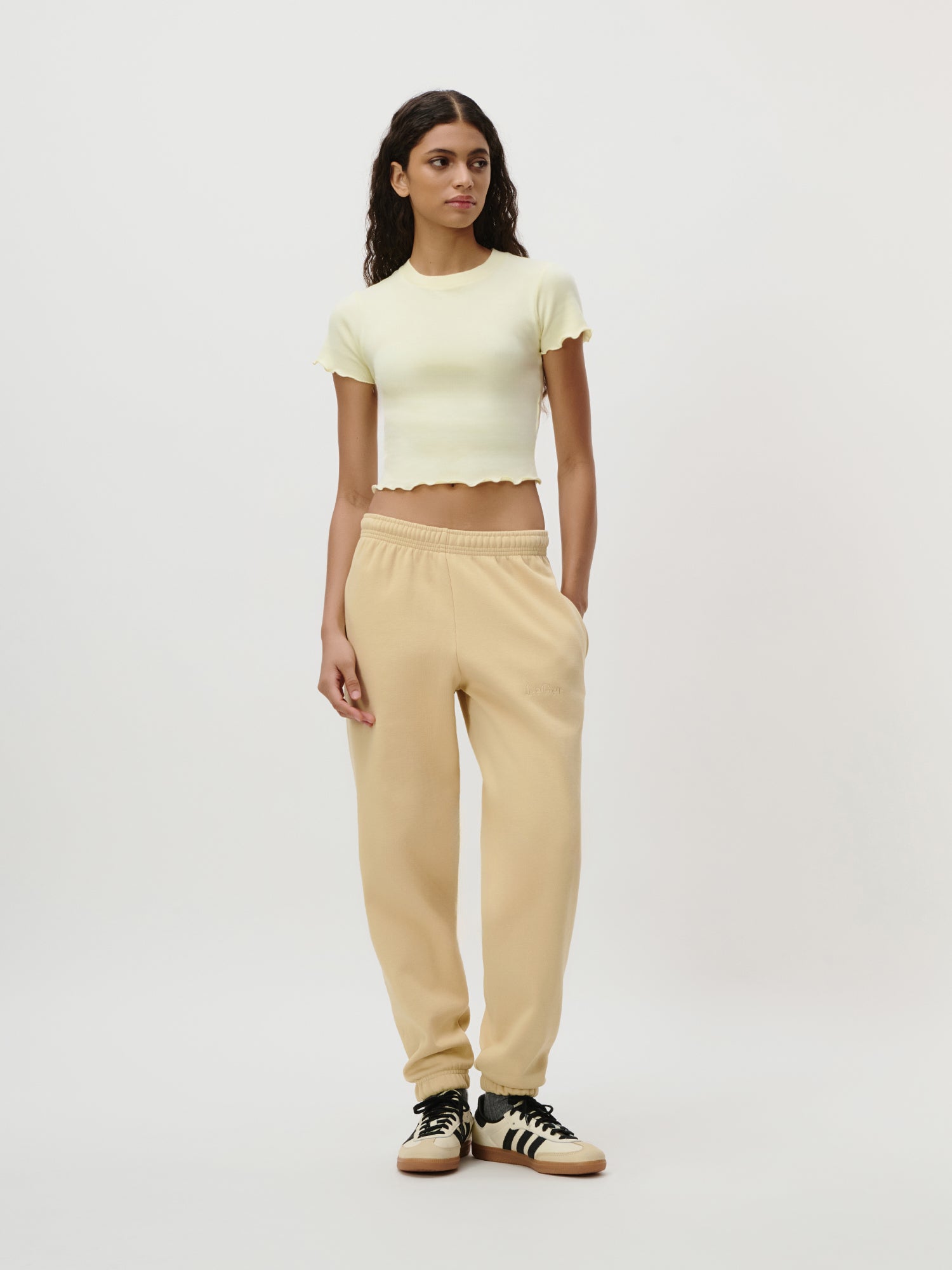 Eine Frau mit langen dunklen Haaren posiert vor einem schlichten Hintergrund und trägt das Teresa T-Shirt von LeGer by Lena Gercke - ein blassgelbes, schmal geschnittenes Oberteil mit gerüschten Kanten - kombiniert mit einer beigen Jogginghose und schwarz-weißen Adidas-Sneakers.