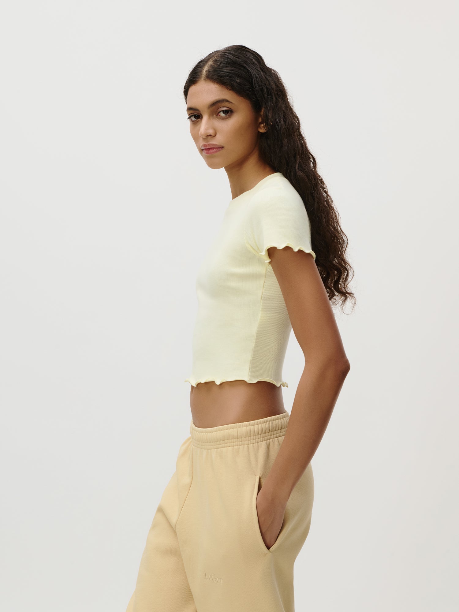 Eine Frau mit langen, gewellten dunklen Haaren steht im Profil vor einem unifarbenen Hintergrund. Sie trägt ein hellgelbes Teresa-T-Shirt von LeGer by Lena Gercke mit gerüschten Rändern und eine beigefarbene Slim-Fit-Jogginghose, die Hände in die Taschen gesteckt.