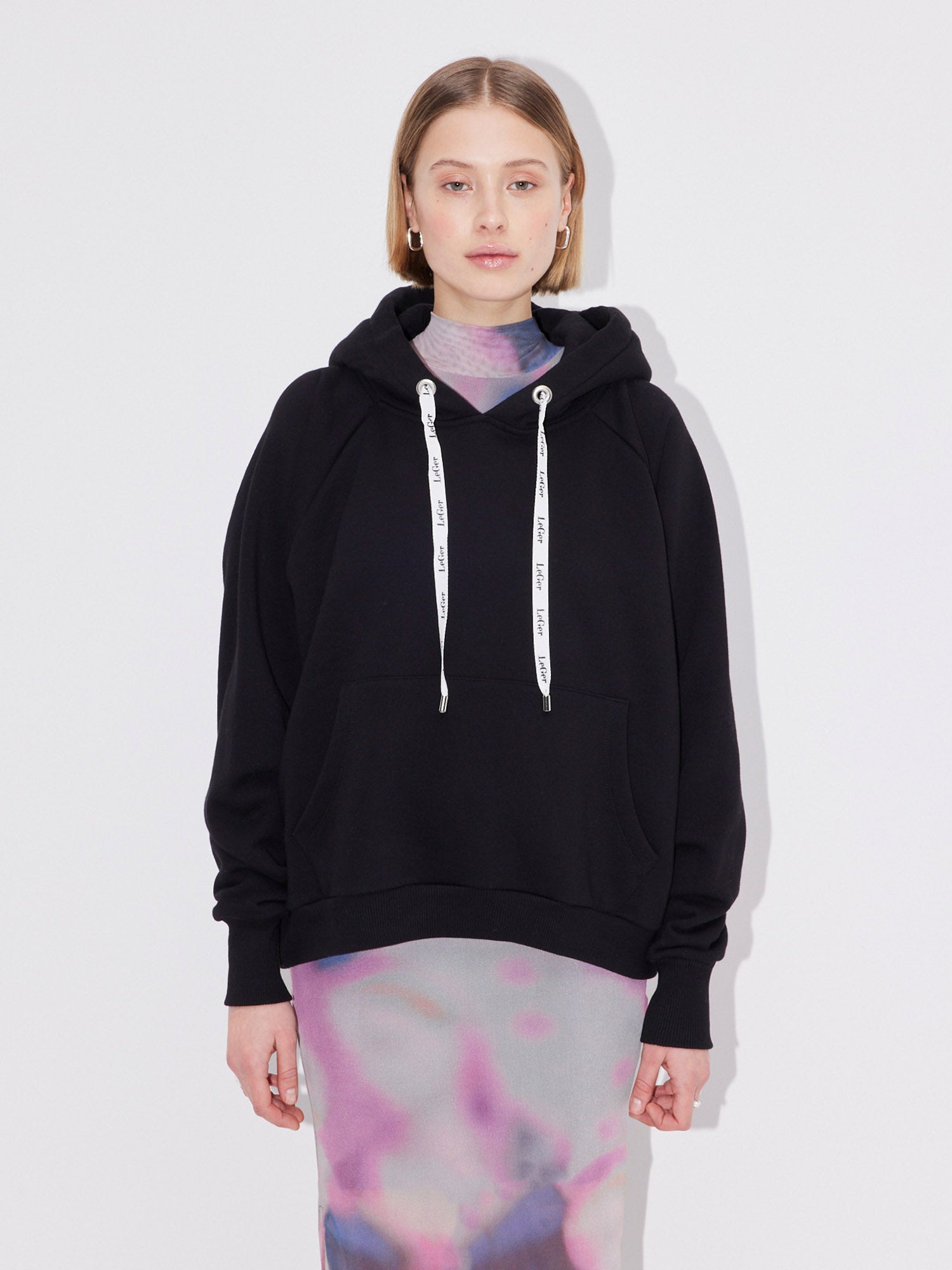 Eine Frau mit schulterlangen blonden Haaren und neutralem Gesichtsausdruck trägt den LeGer by Lena Gercke Hayley Hoodie - einen schwarzen, langärmeligen Kapuzenpullover mit weißem Kordelzug und normaler Passform - über einem pastellfarbenen, halbtransparenten Kleid vor einem schlichten weißen Hintergrund.