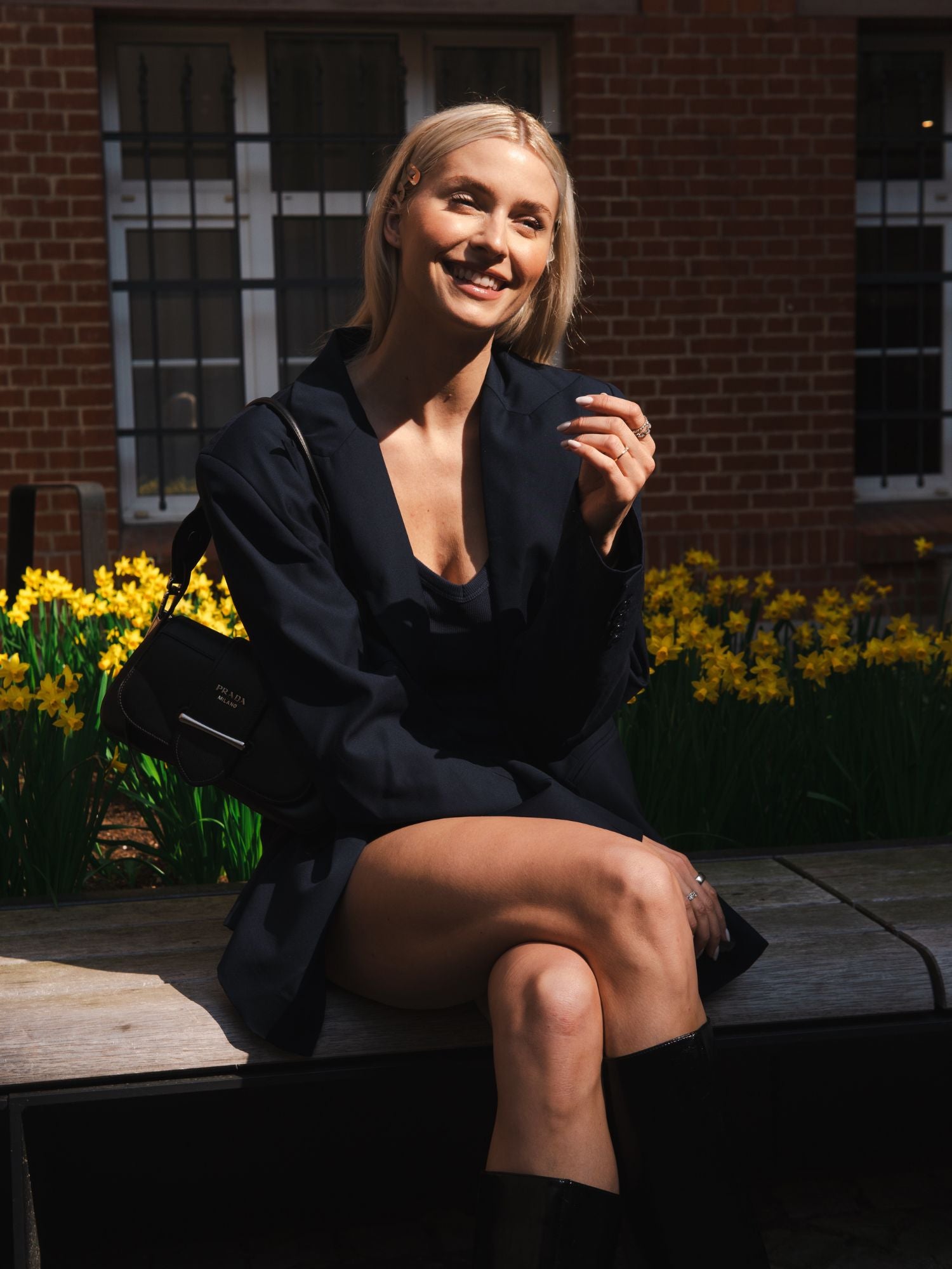 Eine blonde Frau sitzt lächelnd auf einer Bank im Freien. Sie trägt den Blazer „Dita“ von LeGer by Lena Gercke in lockerer Passform, dazu eine schwarze Tasche und kniehohe Stiefel. Hinter ihr sind gelbe Blumen und ein Backsteingebäude mit Fenstern zu sehen.