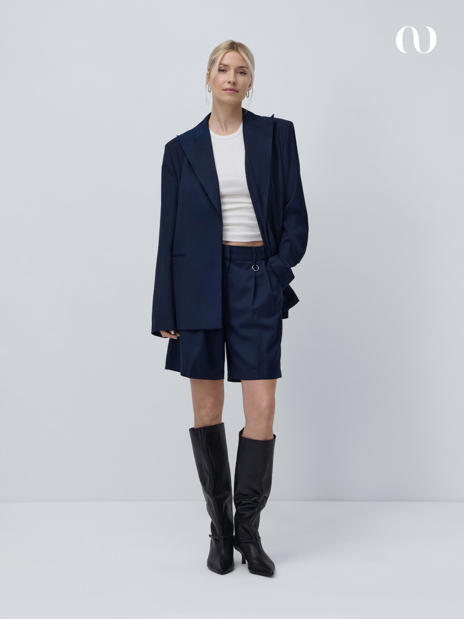 Eine Frau mit hellem Haar und neutralem Gesichtsausdruck trägt die Dita Shorts von LeGer by Lena Gercke, einen marineblauen, locker sitzenden Blazer, ein weißes Crop-Top und schwarze kniehohe Stiefel vor einem schlichten Hintergrund.