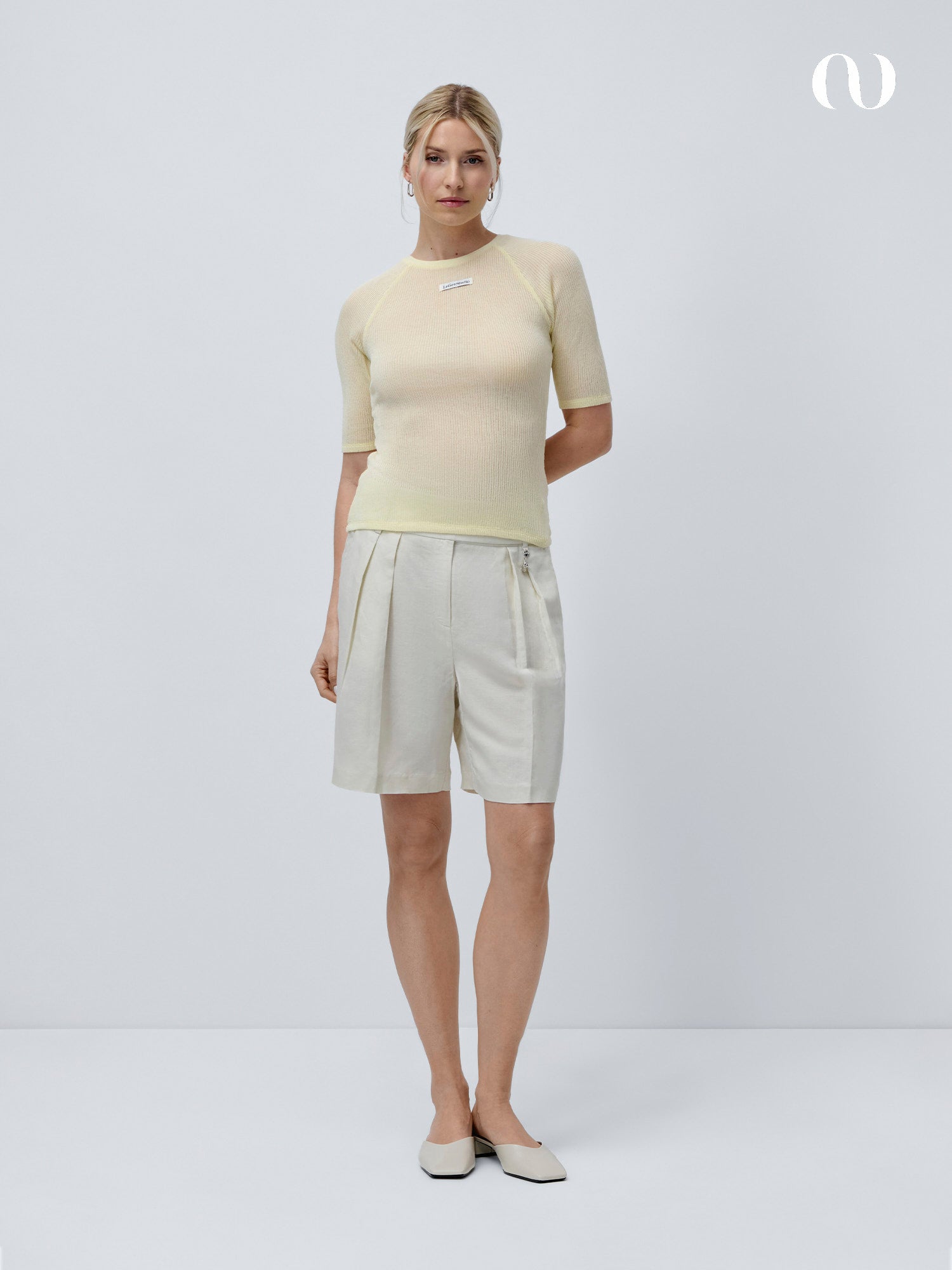 Eine Frau steht vor einem schlichten hellgrauen Hintergrund und trägt das hellgelbe T-Shirt Abby von LeGer by Lena Gercke mit Raglanärmeln, helle Bundfaltenhosen und beigefarbene Schuhe. Ihre Haare sind nach hinten gebunden und ihre Hände befinden sich hinter ihrem Rücken.