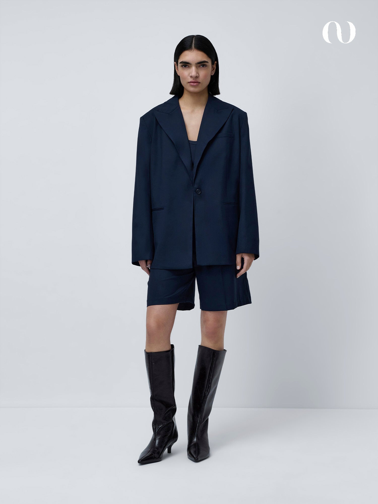 Eine Person mit glattem dunklem Haar trägt einen übergroßen dunkelblauen Blazer, passende Shorts, das schmal geschnittene Kaili Top von LeGer by Lena Gercke mit V-Ausschnitt und schwarze kniehohe Stiefel vor einem schlichten hellgrauen Hintergrund.