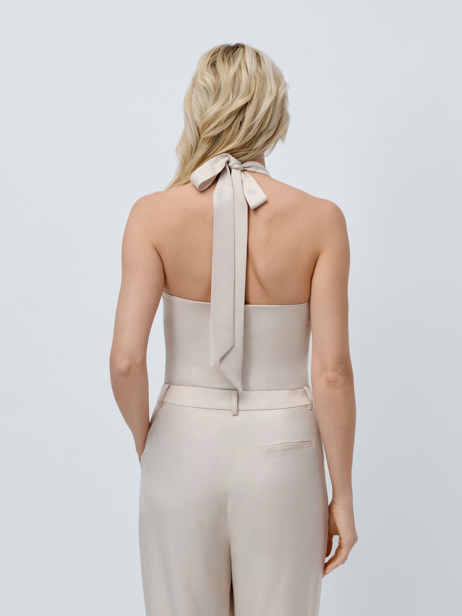 Eine Frau mit blondem, gewelltem Haar ist von hinten zu sehen. Sie trägt das beigefarbene Belinay Top von LeGer by Lena Gercke mit einer langen Krawatte im Nacken, gepaart mit einer passenden, schmal geschnittenen, hoch taillierten Hose. Ihre Hände stecken in den Taschen vor einem schlichten, hellen Hintergrund.