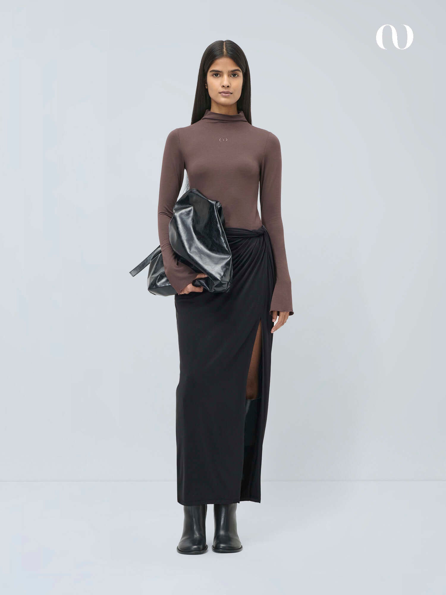 Eine Frau steht vor einem unifarbenen Hintergrund und trägt ein tailliertes braunes langärmeliges Oberteil, den LeGer by Lena Gercke Perle Rock mit oberschenkelhohem Schlitz, schwarze Stiefel und eine große glänzende schwarze Handtasche.