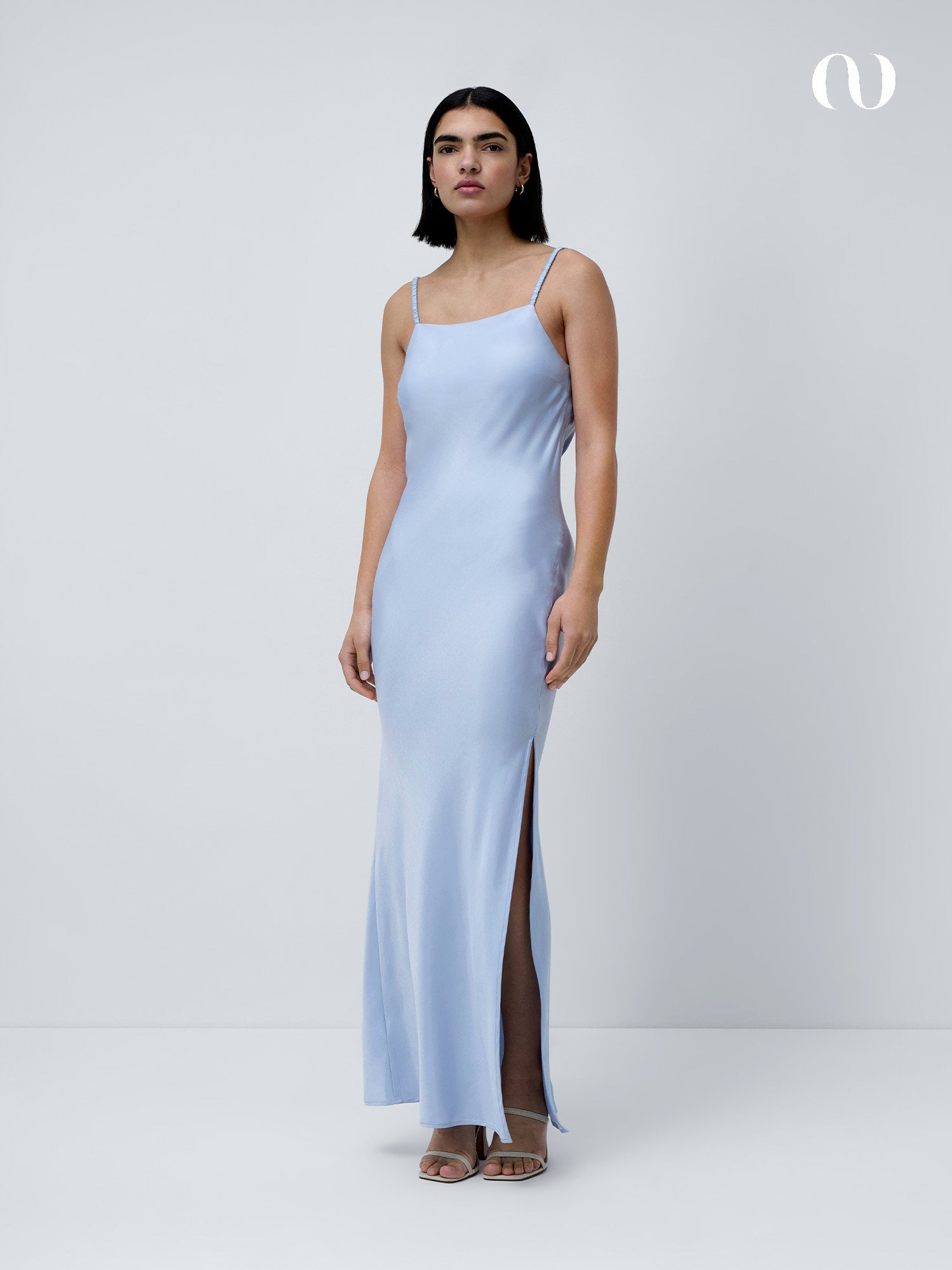 Eine Person mit schulterlangen dunklen Haaren trägt das hellblaue Antonie Dress von LeGer by Lena Gercke, ein schmal geschnittenes Maxikleid mit dünnen Trägern und einem oberschenkelhohen Schlitz, gepaart mit Riemchensandalen, vor einem schlichten weißen Hintergrund.