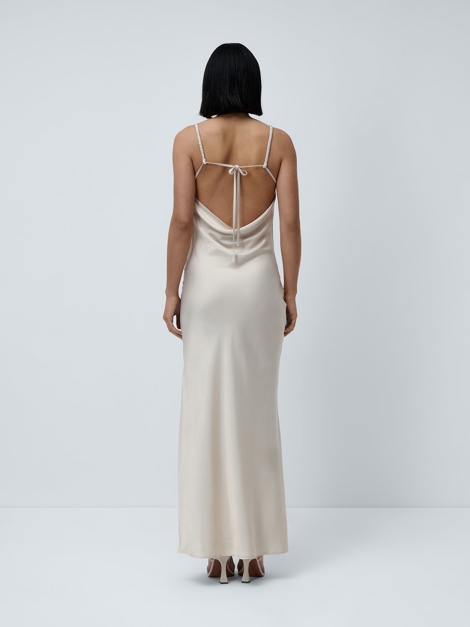 Eine Person mit glattem, dunklem Haar trägt das Kleid „Antonie“ von LeGer by Lena Gercke – ein langes, cremefarbenes Maxikleid aus Satin mit schmalen Trägern, einem geraden Ausschnitt und einem eleganten Cowl-Rückenausschnitt –, während sie vor einem schlichten, hellgrauen Hintergrund steht.