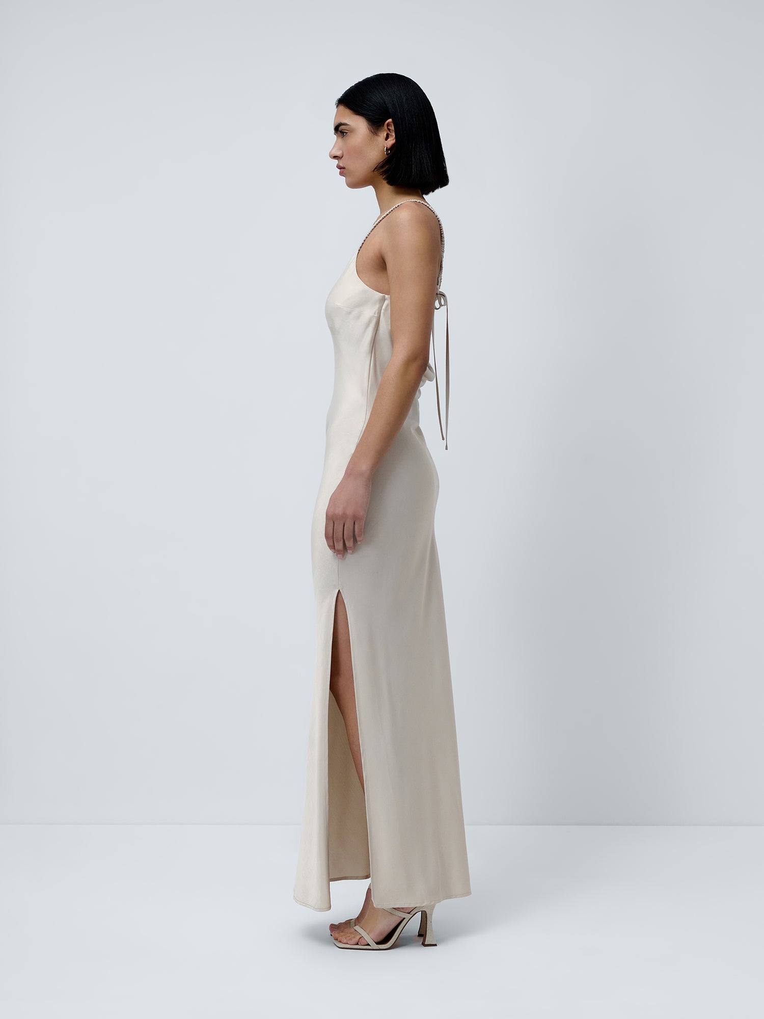 Eine Frau mit glattem, dunklem Haar, die seitlich vor einem schlichten weißen Hintergrund steht, trägt das „Antonie“-Kleid von LeGer by Lena Gercke – ein beiges Maxikleid mit geradem Ausschnitt, schmalen Trägern und einem Seitenschlitz. Sie rundet den Look mit Sandaletten ab.