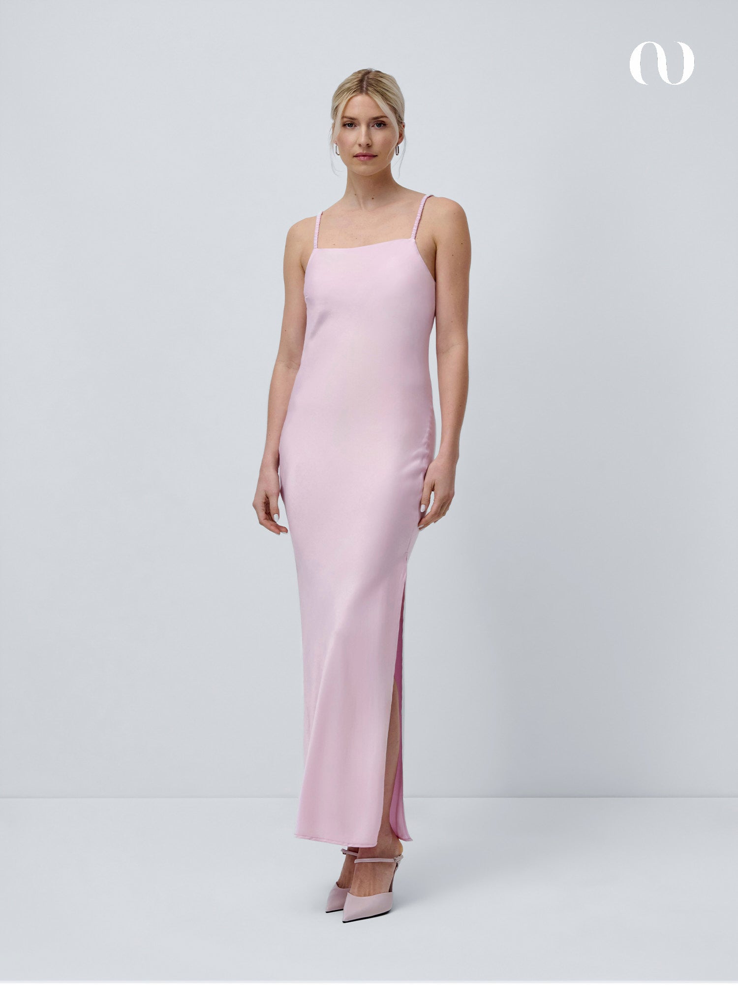 Eine Frau trägt das Antonie-Kleid von LeGer by Lena Gercke in zartem Rosa mit dünnen Trägern und einem Seitenschlitz vor einem weißen Hintergrund. Sie kombiniert das schmal geschnittene Kleid mit hellrosa Absätzen und trägt ihr blondes Haar zu einer Hochsteckfrisur.