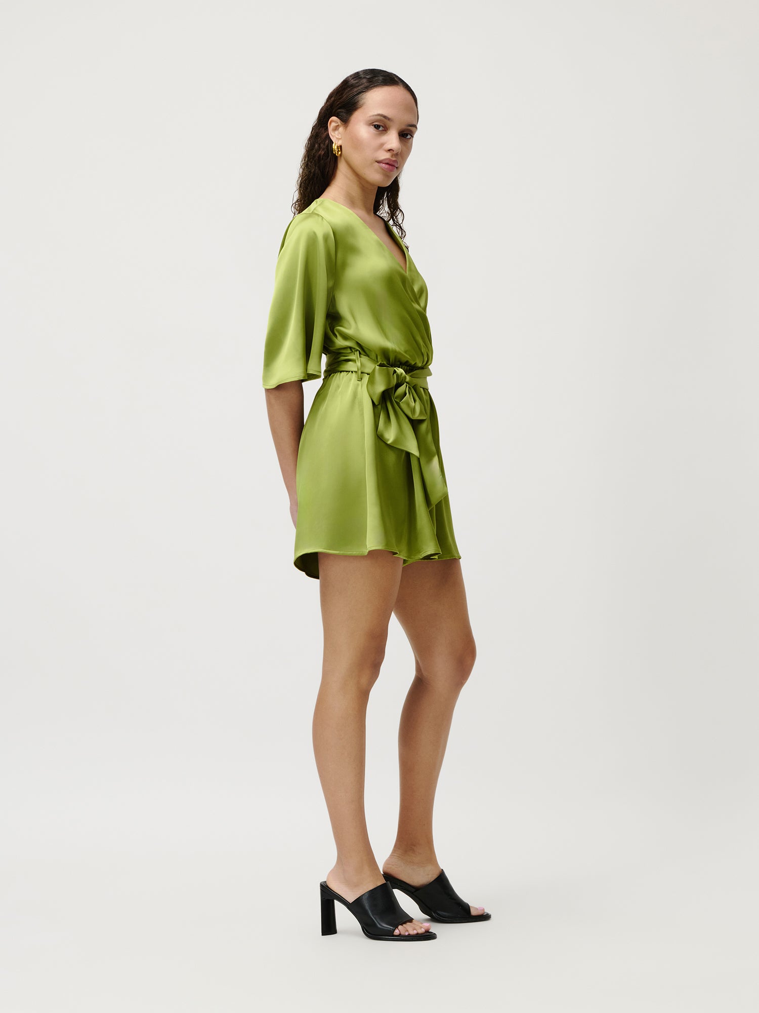 Eine Frau steht seitlich und trägt den Alexis-Overall von LeGer by Lena Gercke mit schwarzen Pantoletten mit Absatz vor einem schlichten weißen Hintergrund und präsentiert mühelose Mode.