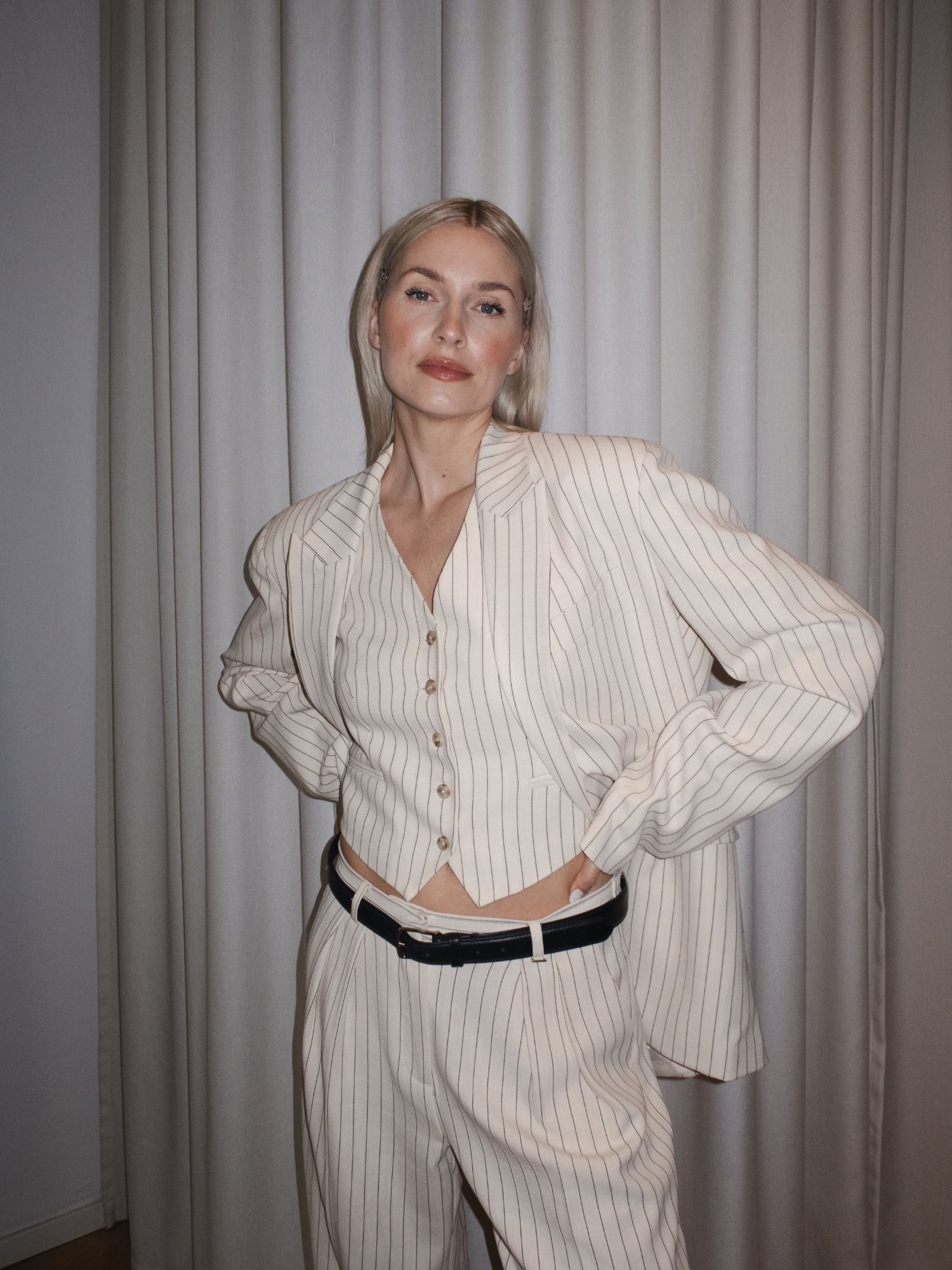 Eine Frau steht vor hellen Vorhängen und trägt den beigen Elaine Blazer von LeGer by Lena Gercke in Oversized-Passform mit extralangen Ärmeln, gepaart mit einer passenden Weste und einer Hose, die mit einem schwarzen Gürtel abgeschlossen wird.