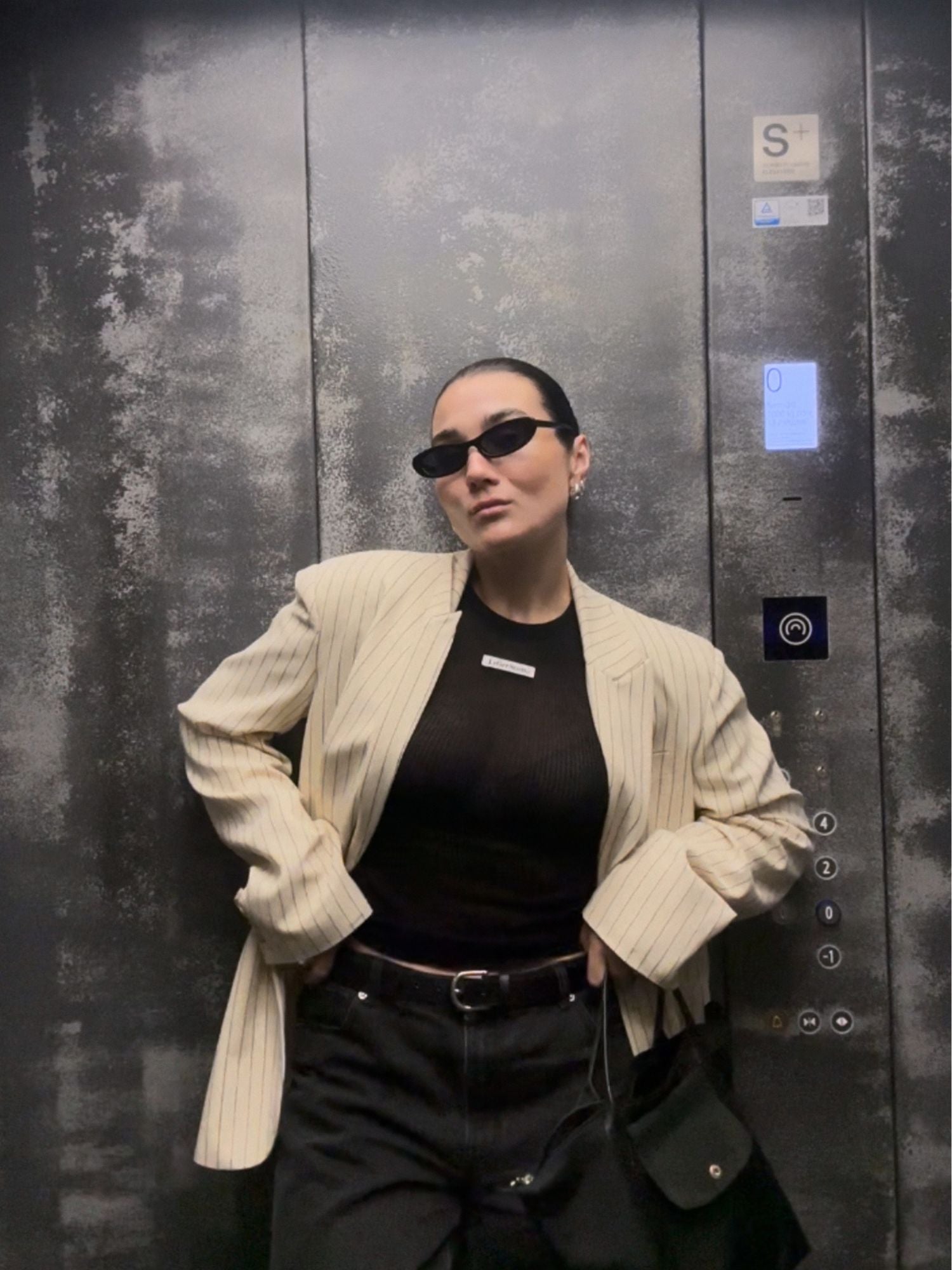 Eine Frau mit schwarzer Sonnenbrille und einem übergroßen beigen „Elaine“-Blazer von LeGer by Lena Gercke posiert selbstbewusst vor einer metallischen Aufzugswand mit sichtbaren Knöpfen und einem kleinen Bildschirm.