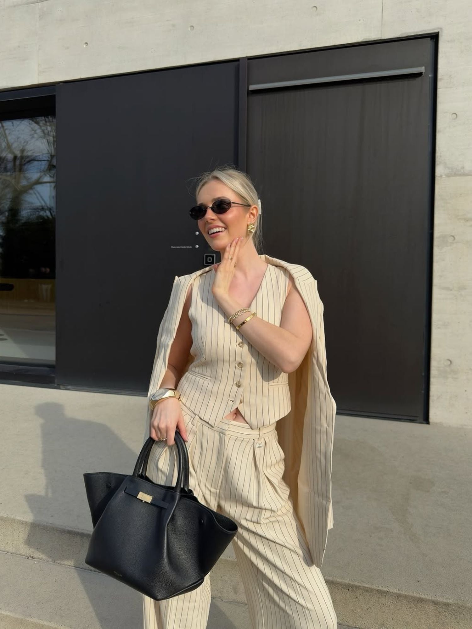 Eine Frau, die die LeGer by Lena Gercke Elaine Vest mit verstellbarer Taille und den passenden Nadelstreifenblazer trägt, lächelt, winkt und hält eine schwarze Handtasche. Sie trägt eine Sonnenbrille und Goldschmuck vor einem modernen Betongebäude.