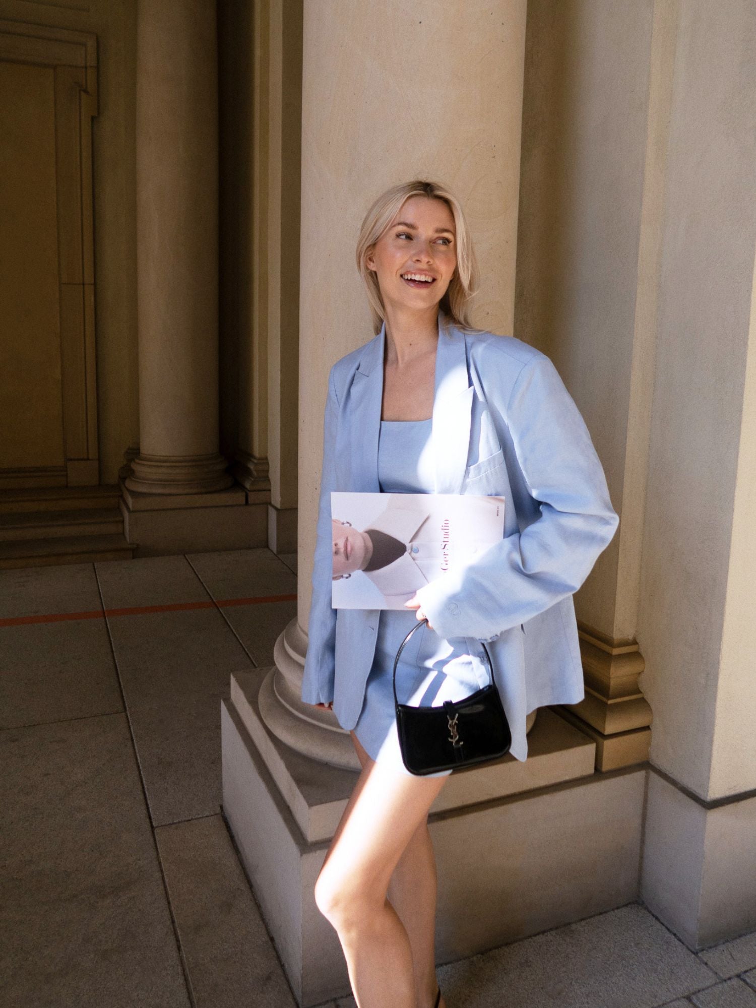 Eine Frau im Majella-Rock von LeGer by Lena Gercke steht in der Nähe von Steinsäulen, hält eine Zeitschrift und eine kleine schwarze Handtasche in der Hand, lächelt und schaut zur Seite.