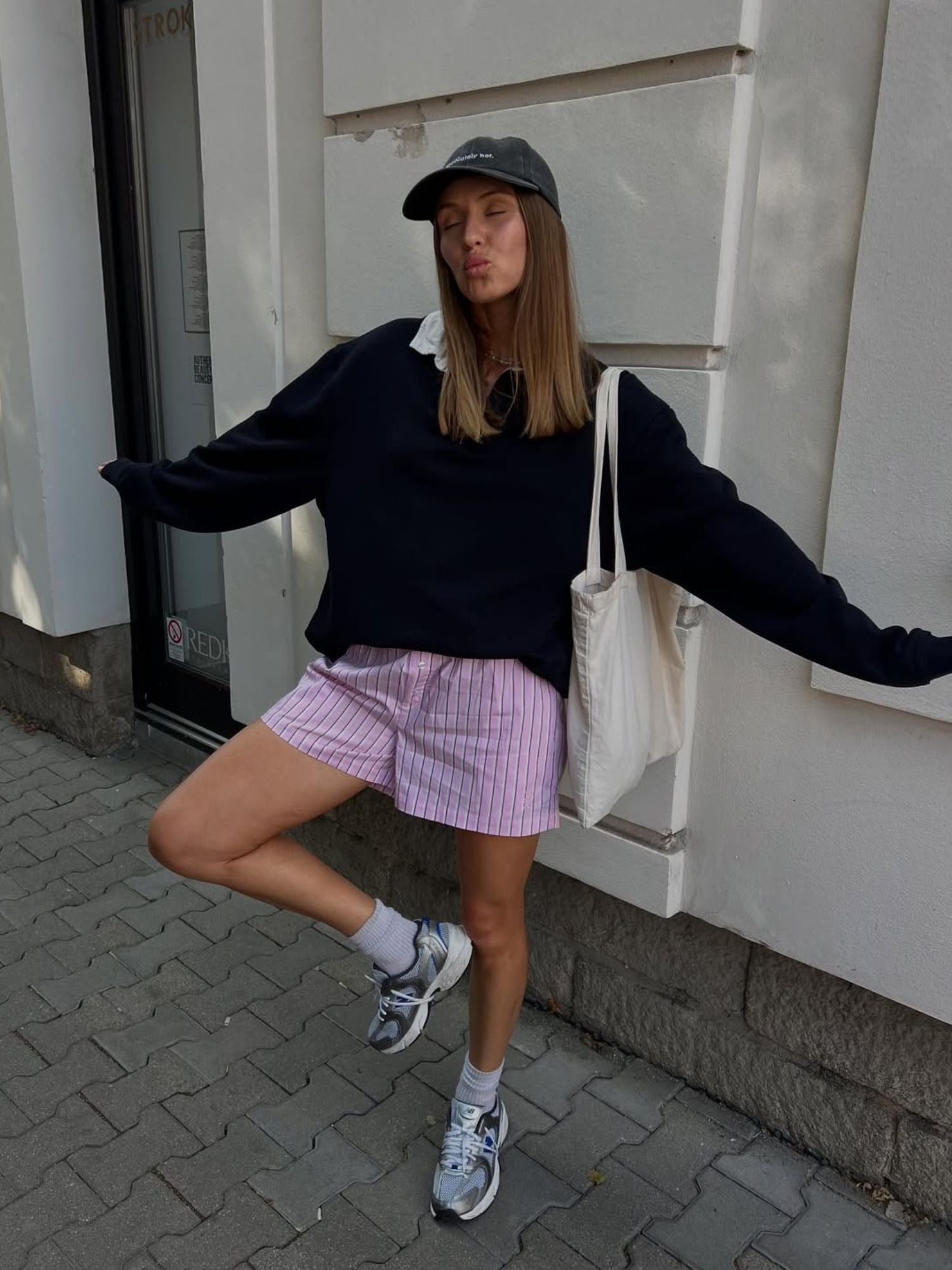 Eine junge Frau in Größe 36 lächelt verspielt auf einem Bürgersteig und modelliert die Farine Shorts von LeGer by Lena Gercke mit einem schwarzen Pullover, Turnschuhen, einer schwarzen Mütze und einer weißen Tragetasche. Sie posiert auf einem Bein, mit zusammengekniffenen Lippen und geschlossenen Augen vor einer weißen Wand.