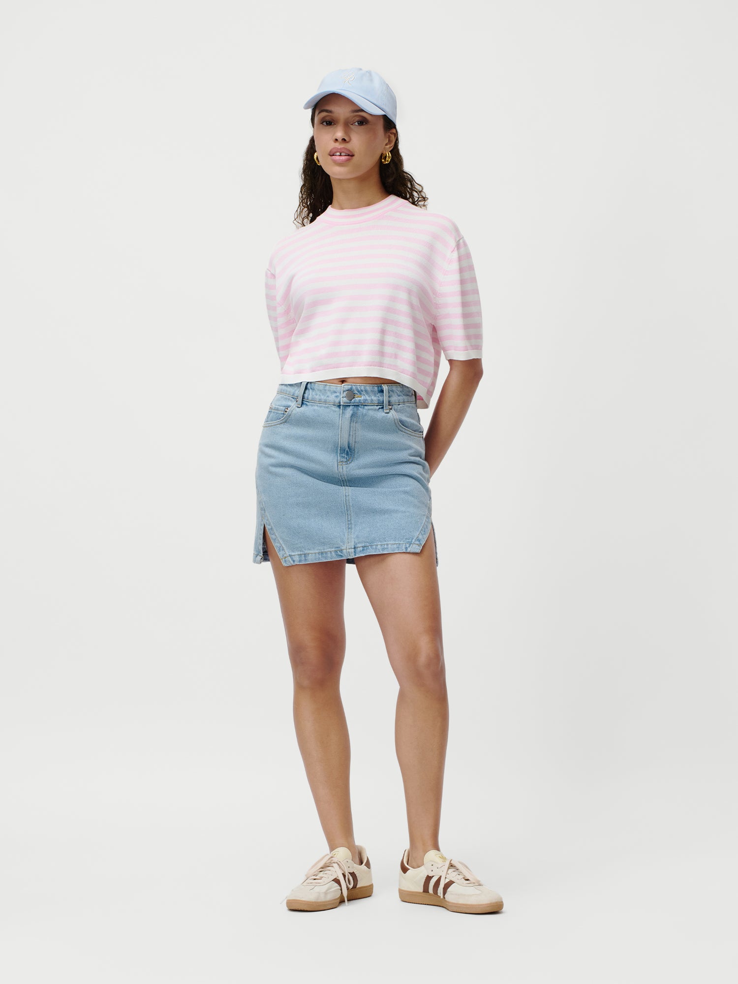 Eine Frau trägt eine hellblaue Baseballkappe, ein rosa-weiß gestreiftes T-Shirt, den Daphne Rock von LeGer by Lena Gercke im Denim-Stil mit Seitenschlitzen und beigefarbene Turnschuhe vor einem schlichten weißen Hintergrund.