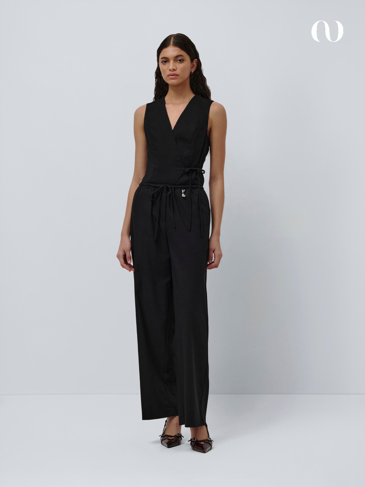 Eine Frau steht vor einem hellgrauen Hintergrund und trägt die Emelie Pants von LeGer by Lena Gercke - ärmellos, schwarz, mittlere Taille, lockere Passform mit weiten Beinen - kombiniert mit schwarzen spitzen Schuhen.