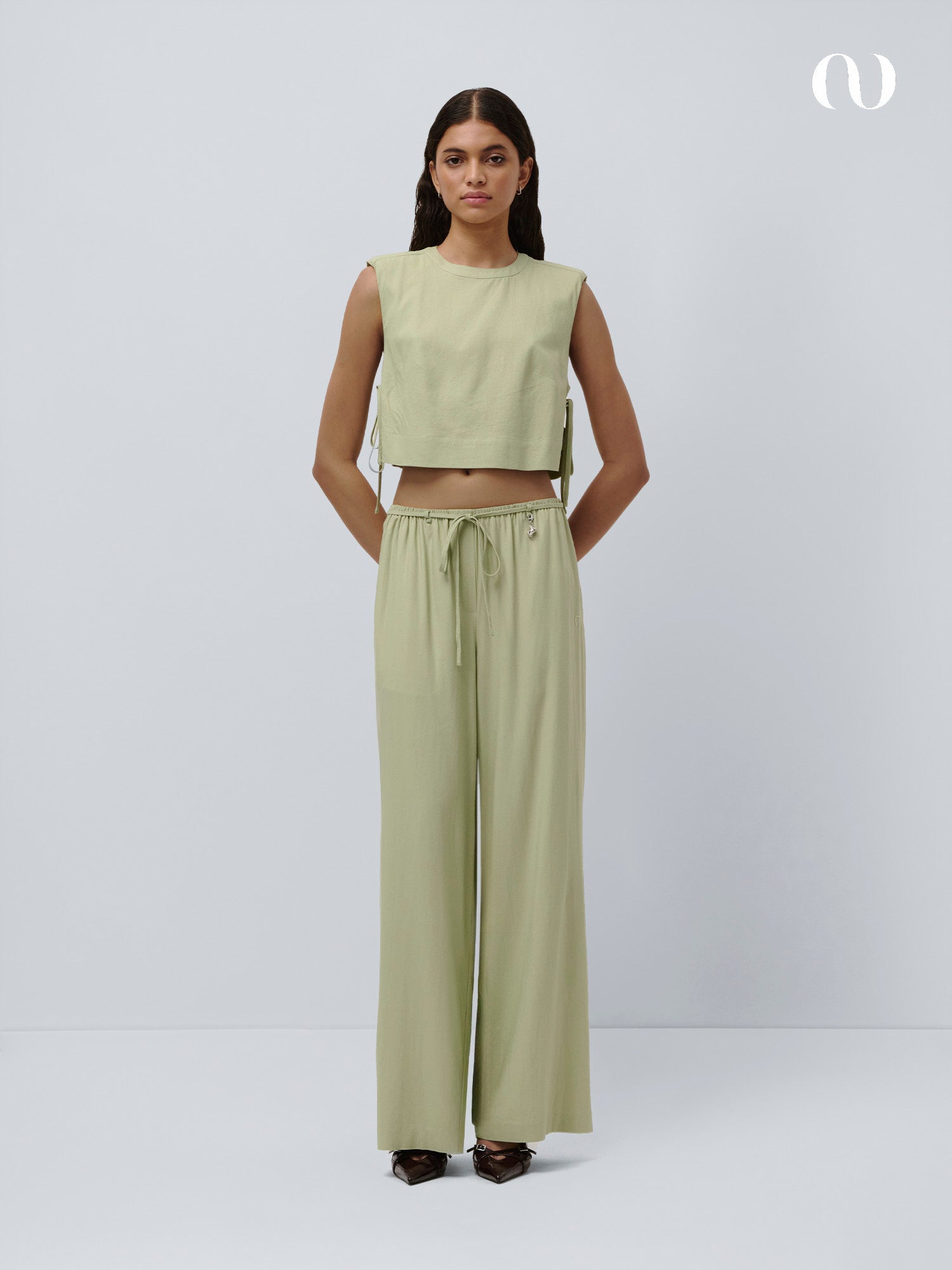 Eine Frau steht vor einem schlichten Hintergrund, trägt ein ärmelloses hellgrünes Crop-Top und die Emelie Pants von LeGer by Lena Gercke - eine locker sitzende Hose im Paperbag-Stil mit Kordelzug in der Taille - und blickt mit den Händen in den Taschen und dunklen spitzen Schuhen nach vorn.
