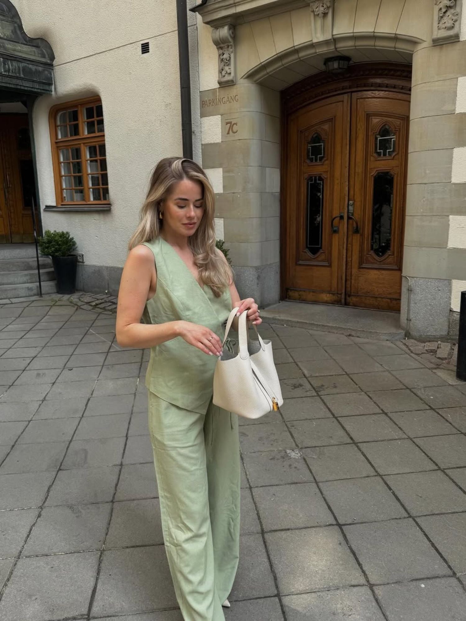 Eine Frau mit langen blonden Haaren, die das Emelie Top von LeGer by Lena Gercke und eine dazu passende Hose in zartem Grün trägt, steht auf einem gepflasterten Gehweg vor einem Gebäude und hält eine weiße Handtasche in der Hand, auf die sie hinunterblickt.