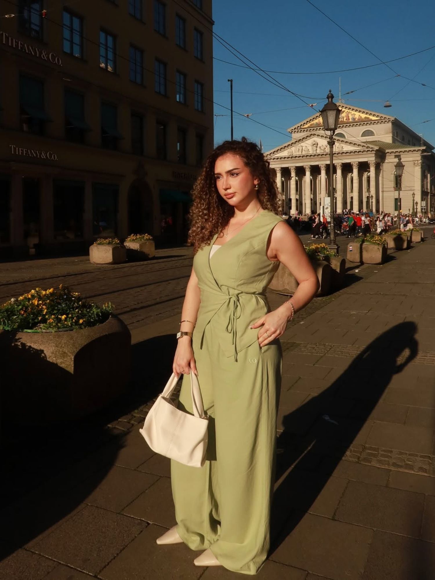Eine Frau trägt das ärmellose Emelie Top in sanftem Grün von LeGer by Lena Gercke mit Wickel-Detail, posiert auf einer sonnigen Stadtstraße vor historischen Gebäuden und hält eine weiße Tasche im warmen Licht.