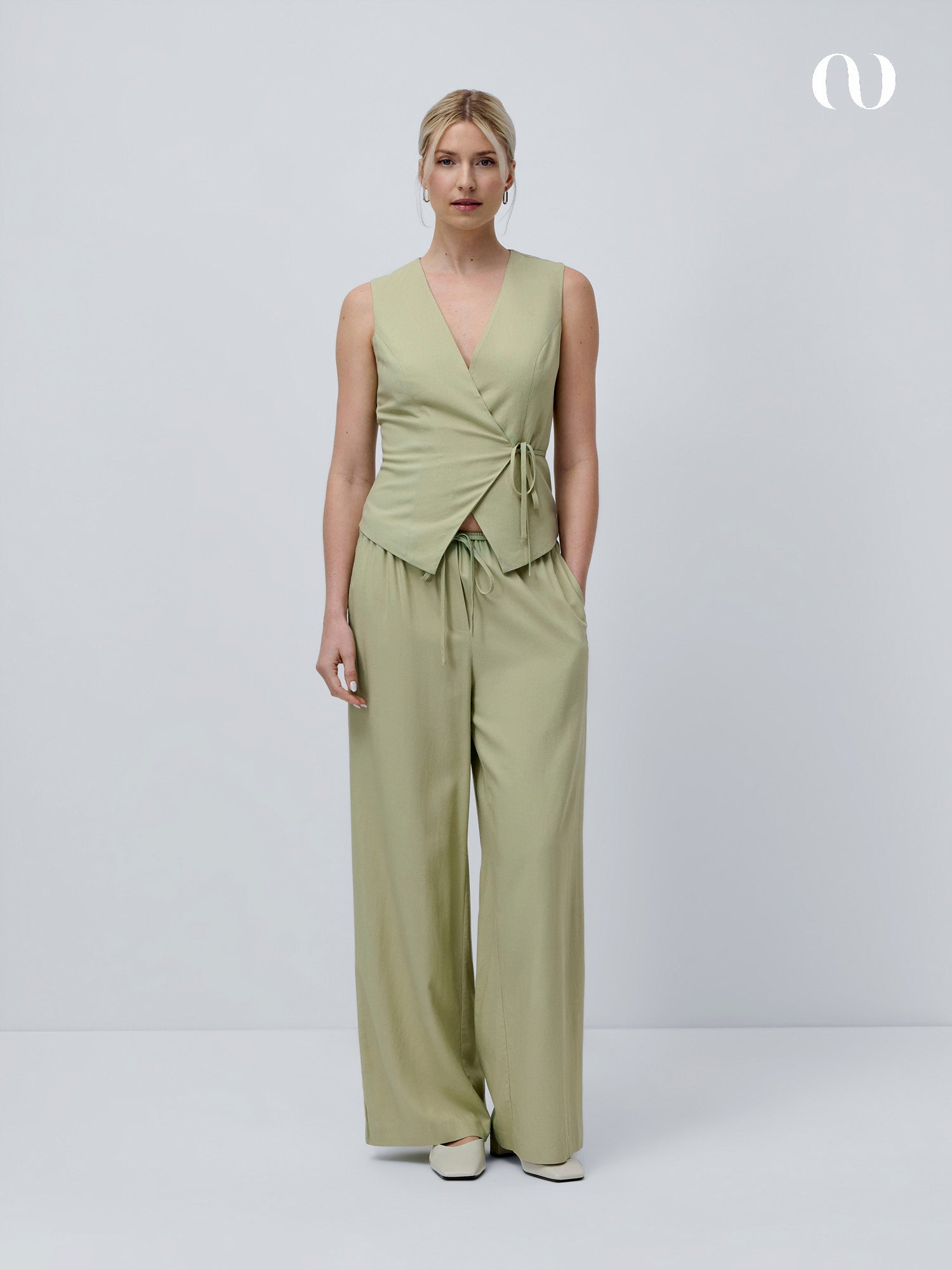 Eine Frau steht vor einem hellgrauen Hintergrund und trägt das hellgrüne Oberteil Emelie mit Wickeloptik und Bindegürtel von LeGer by Lena Gercke, gepaart mit einer passenden Hose mit weitem Bein. Ihr helles Haar ist zurückgebunden, und sie trägt weiße Schuhe.